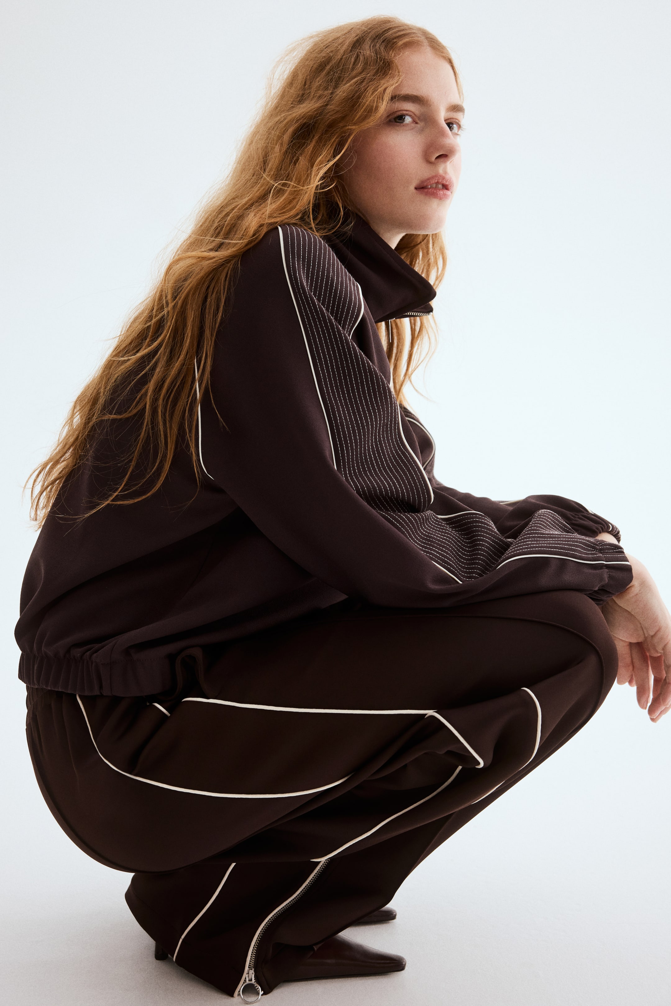 View larger image: A dark brown tracksuit is displayed from a side-back angle, featuring white piping along the sleeves and outer leg seams, with additional white stitched detailing on the upper arms. The jacket showcases a high collar and an elasticated hem, while the trousers feature zips at the ankles.