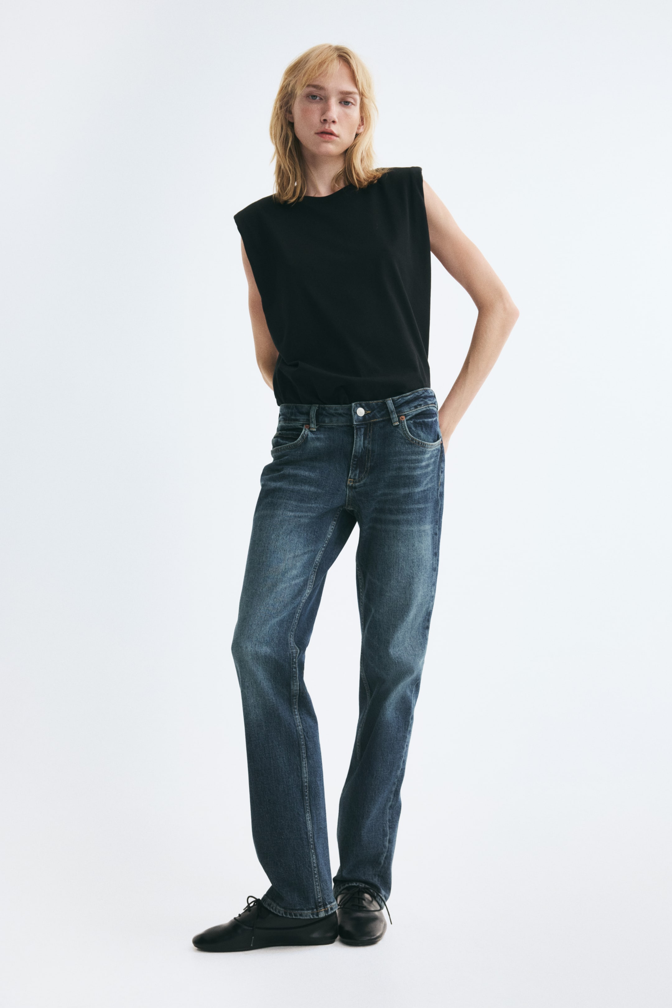 Größeres Bild anzeigen: Blaue Five-Pocket-Jeans aus Denim mit niedriger Leibhöhe, Used-Effekten an den Oberschenkeln und geradem Bein, getragen vom Model. Die Jeans sind mit einem in den Bund gesteckten schwarzen ärmellosen Top und schwarzen flachen Schuhen kombiniert.