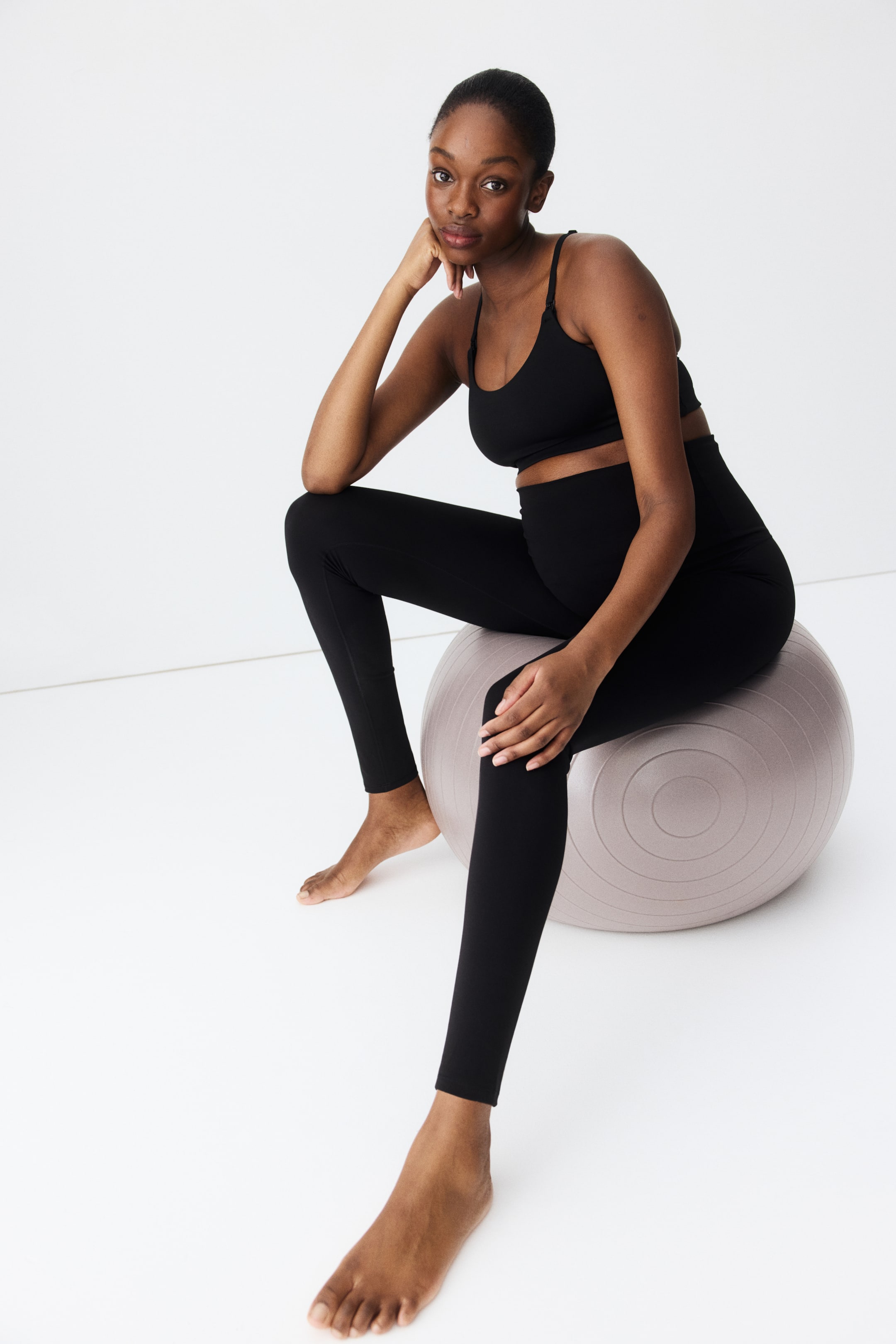 Ampliar la imagen: Un conjunto de ropa de maternidad deportiva negra presenta unas mallas de cintura alta que cubren el vientre y un sujetador deportivo con tirantes delgados ajustables y broches visibles para lactancia.