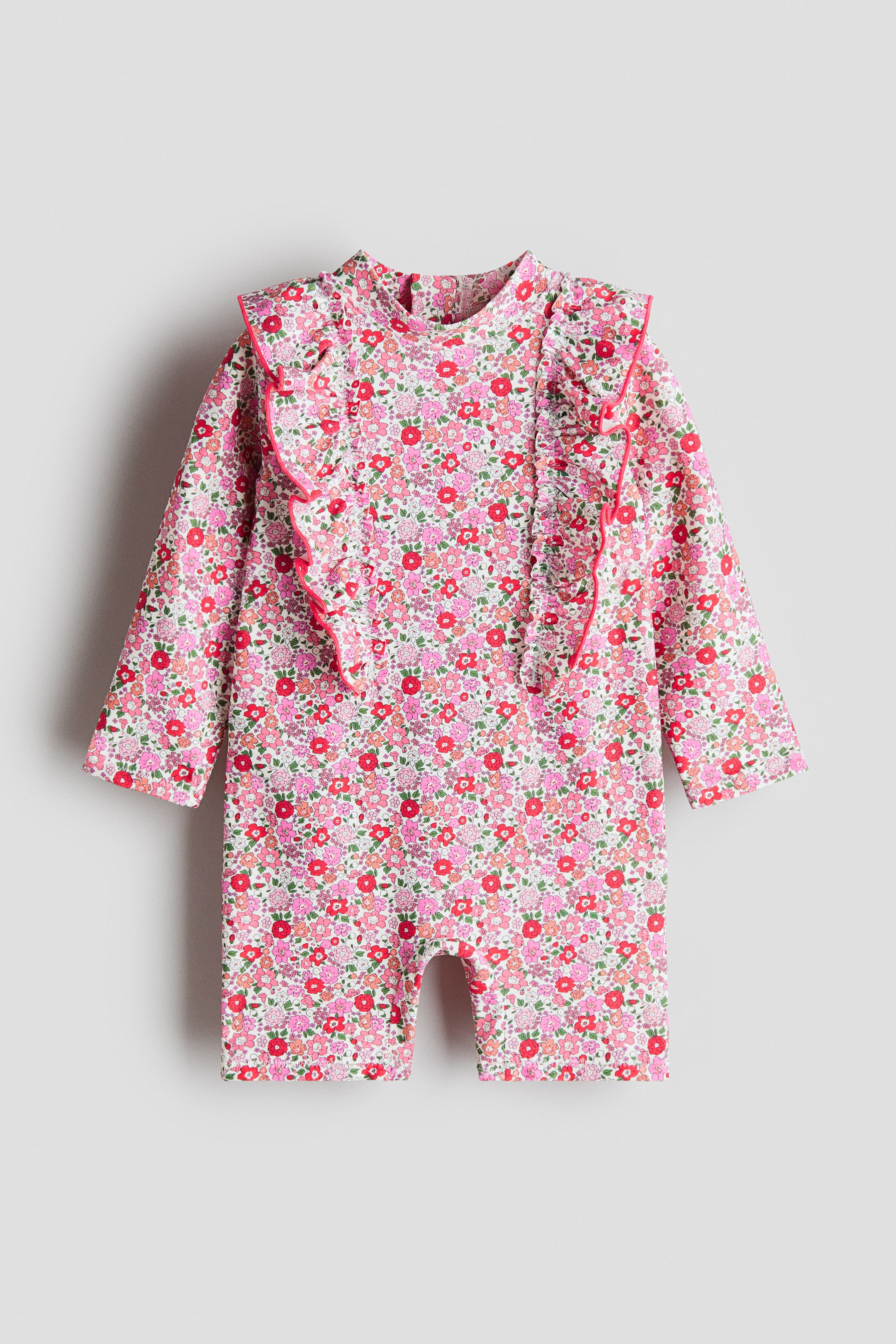 View larger image: The front of the swimsuit displays a pink, red, and green floral print on a white textured fabric. It features a small stand collar, long sleeves, and short legs, with pink-edged frills running down the front and extending over the shoulders.