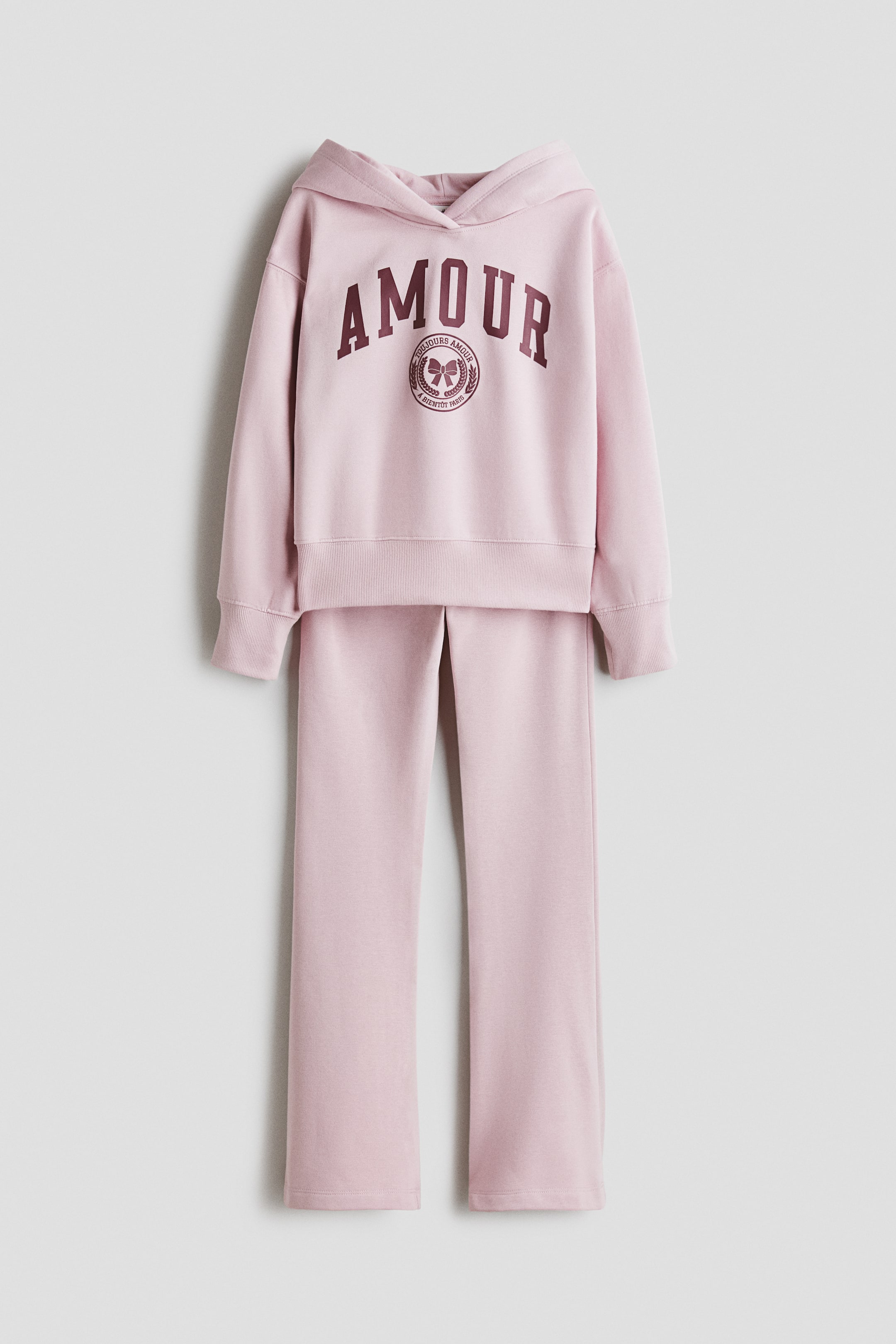 View larger image: A light pink two-piece tracksuit is hanging, shown from the front, consisting of a hoodie and straight-leg sweatpants. The hoodie features a matching hood, long sleeves with ribbed cuffs, a ribbed hem, and a dark red "AMOUR" graphic arched over a circular emblem on the chest.