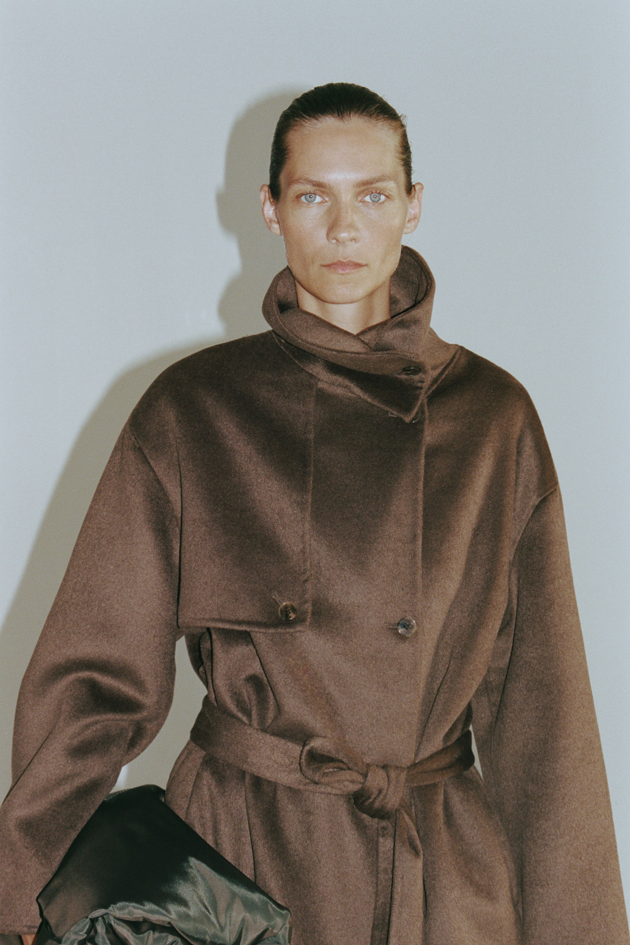 View larger image: The model is wearing a dark brown, felted wool-blend coat featuring a high buttoned funnel collar, a buttoned storm flap across the chest and a matching tie belt at the waist. The model is holding a dark green, satin-like bag tucked under one arm.