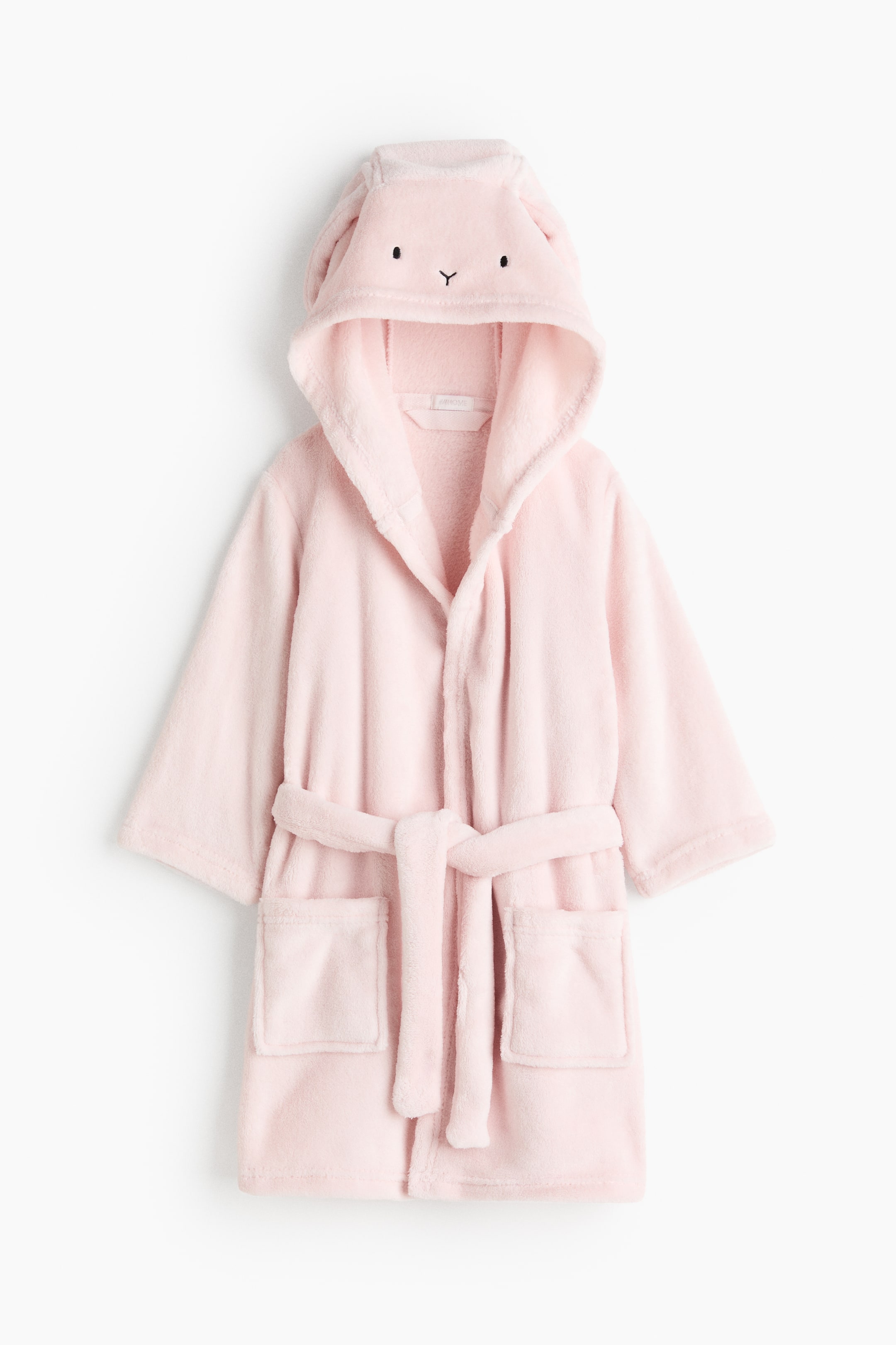 拡大画像を表示: 淡いピンクのふんわりとしたバスローブを正面から撮影。フードには刺繍で施したうさぎの顔立ちと、付け耳の垂れ耳デザインがあしらわれています。ローブは、長くゆったりとした袖、ウエストの共布ベルト、前面のパッチポケット2つを備えています。