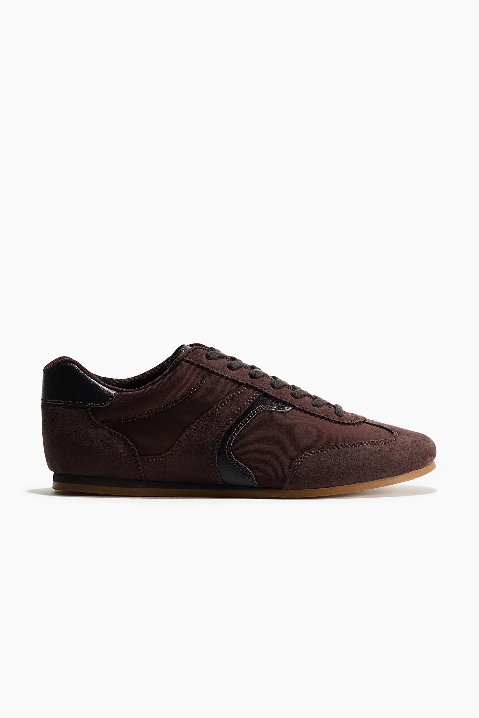 A dark brown low-top sneaker is facing right in side profile, featuring a combination of textile and suede panels with front lacing and a padded collar. The shoe showcases a dark leather heel tab and a curved side accent above a thin gum sole.