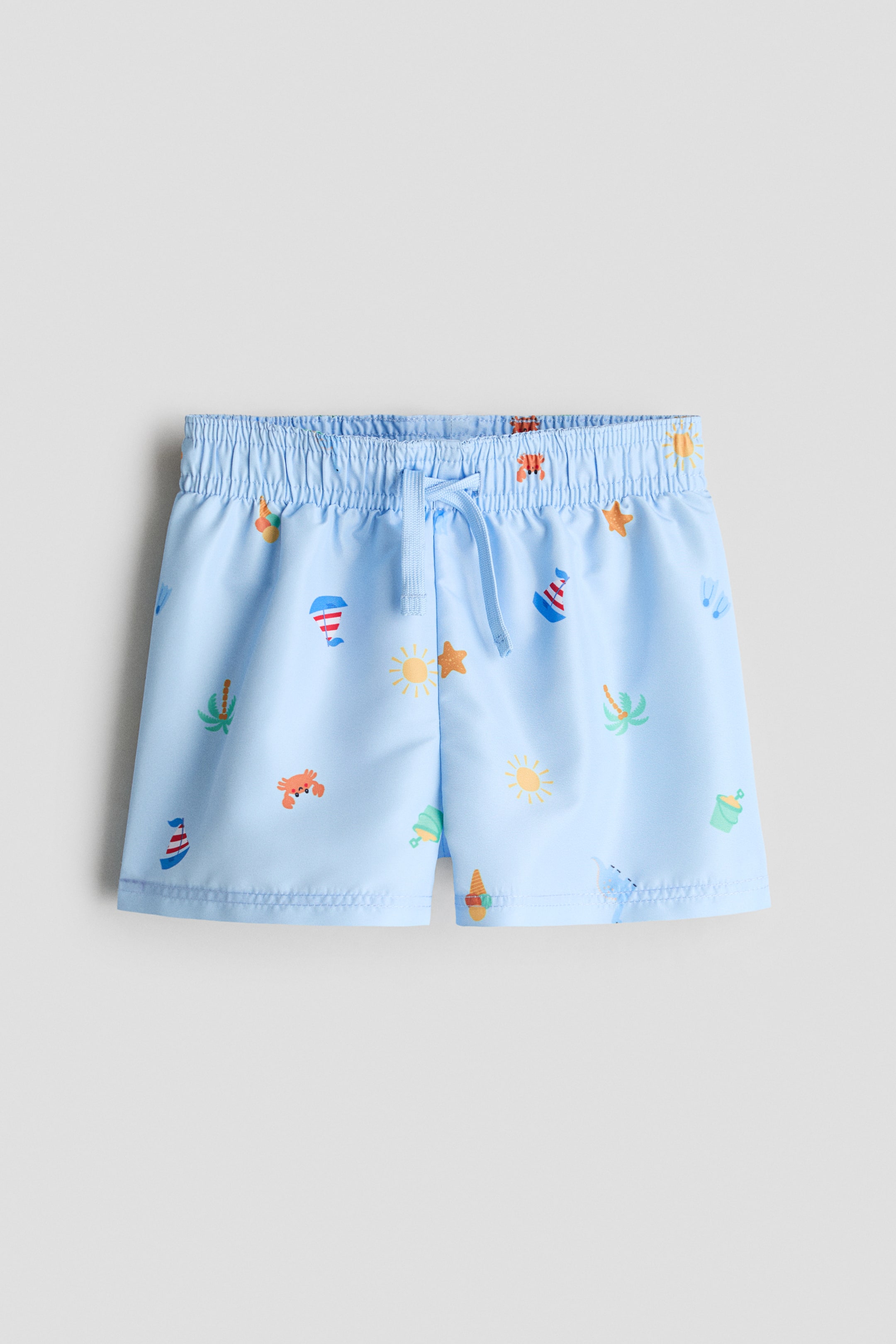 Visualizza immagine più grande: Pantaloncini da bagno azzurri, presentati frontalmente, caratterizzati da un motivo all-over di illustrazioni colorate a tema spiaggia come soli, granchi e barche a vela. Sono progettati con una vita elasticizzata con coulisse, assicurata con un laccio azzurro, e un dettaglio di patta finta cucita sul davanti.