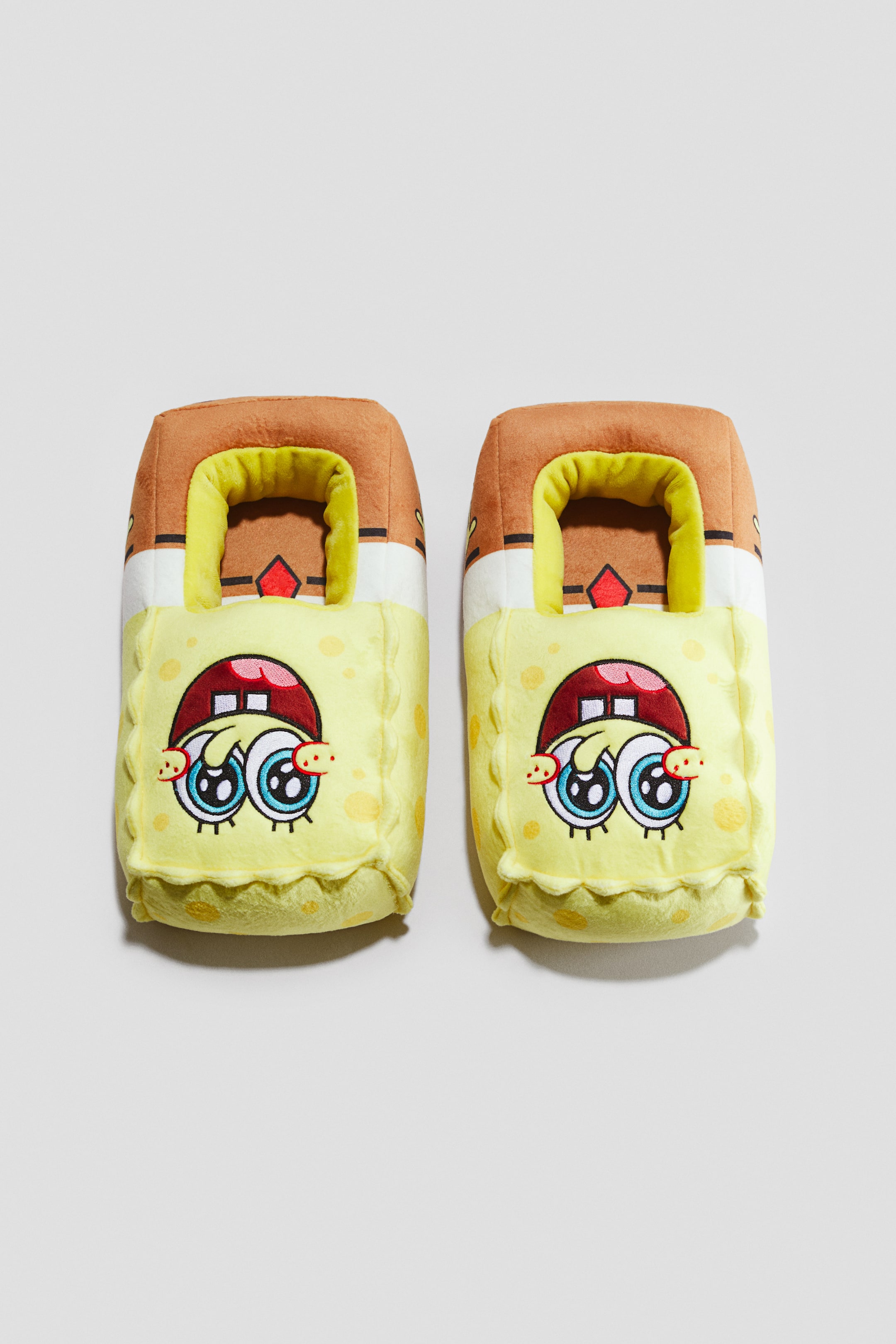 Ampliar la imagen: Dos pantuflas de Bob Esponja de peluche están colocadas una junto a la otra, de frente. Cada pantufla presenta una parte superior café con una banda blanca y detalle de lazo rojo, mientras que la parte frontal amarilla muestra ojos bordados, mejillas sonrosadas, una boca abierta con dientes y una lengua roja, sutiles lunares y un ribete ondulado.
