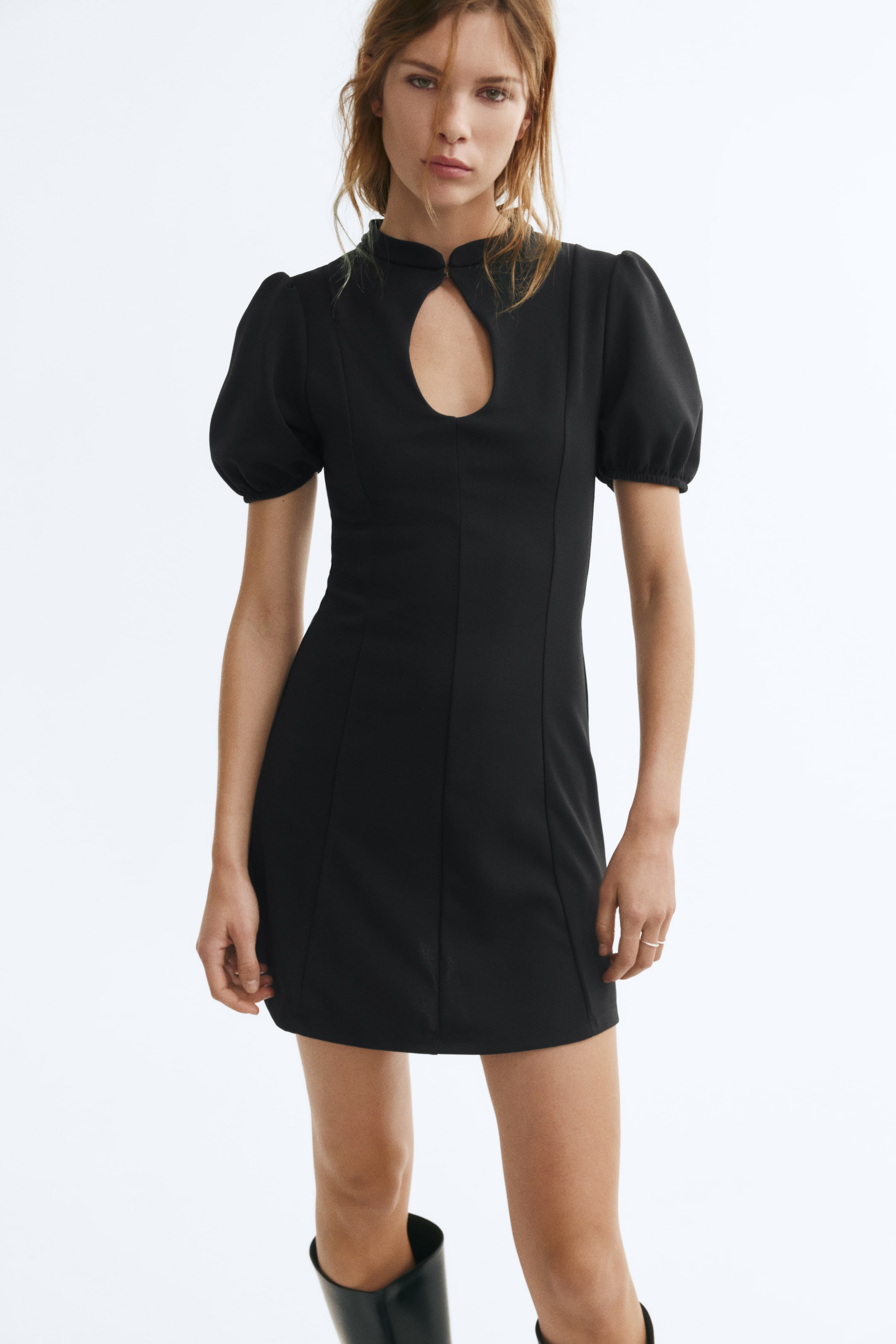 View larger image: A black mini dress is being worn by the model, featuring a fitted bodice, a high neckline with a front keyhole opening, short puff sleeves, and prominent vertical seams down the front. The dress is styled with black knee-high boots.