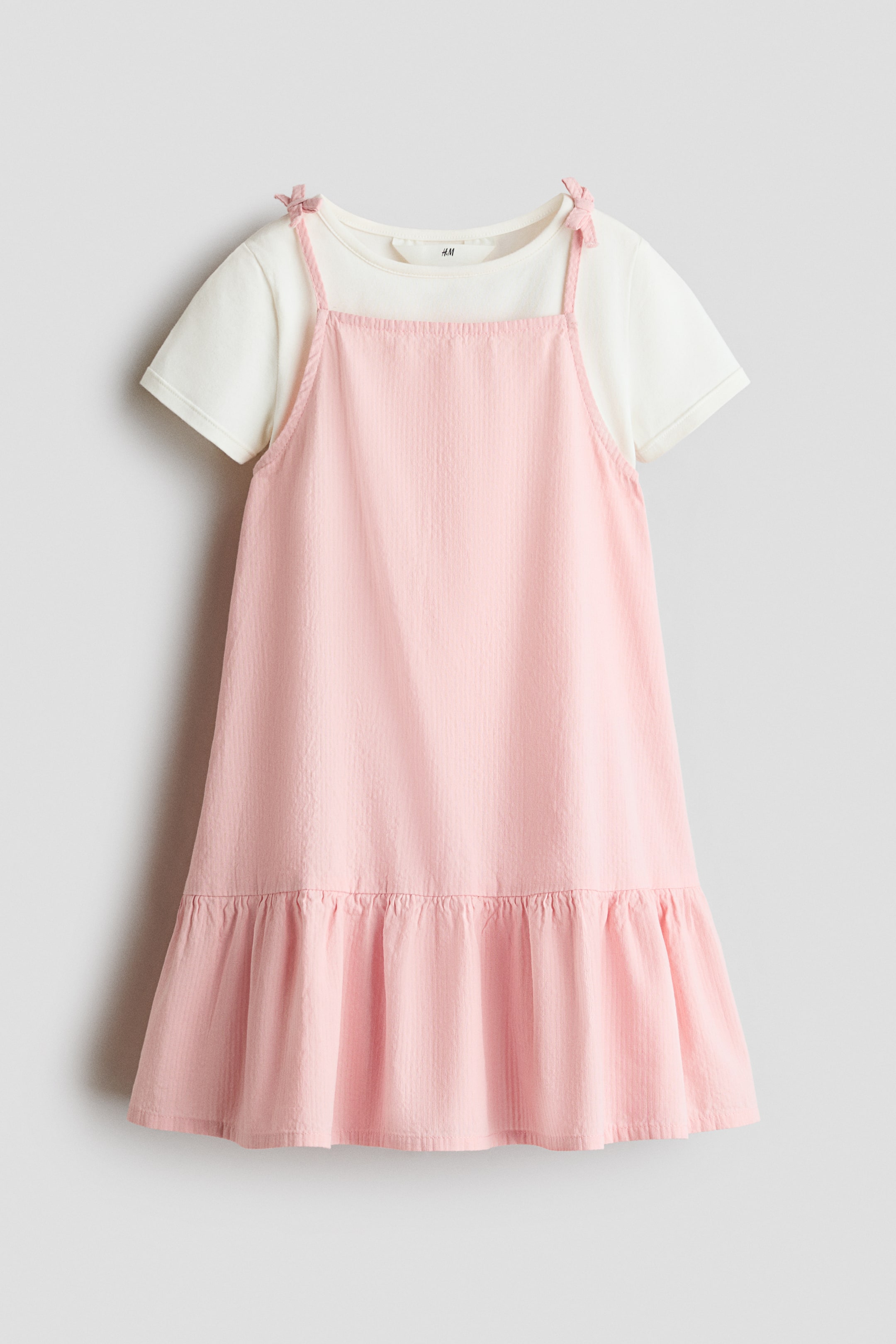 Voir une image plus grande: Une robe tablier texturée rose est superposée sur un t-shirt crème à manches courtes, présentant l’ensemble de face, avec la robe dotée d’un col carré, de bretelles à nouer aux épaules et d’un ourlet à volants.