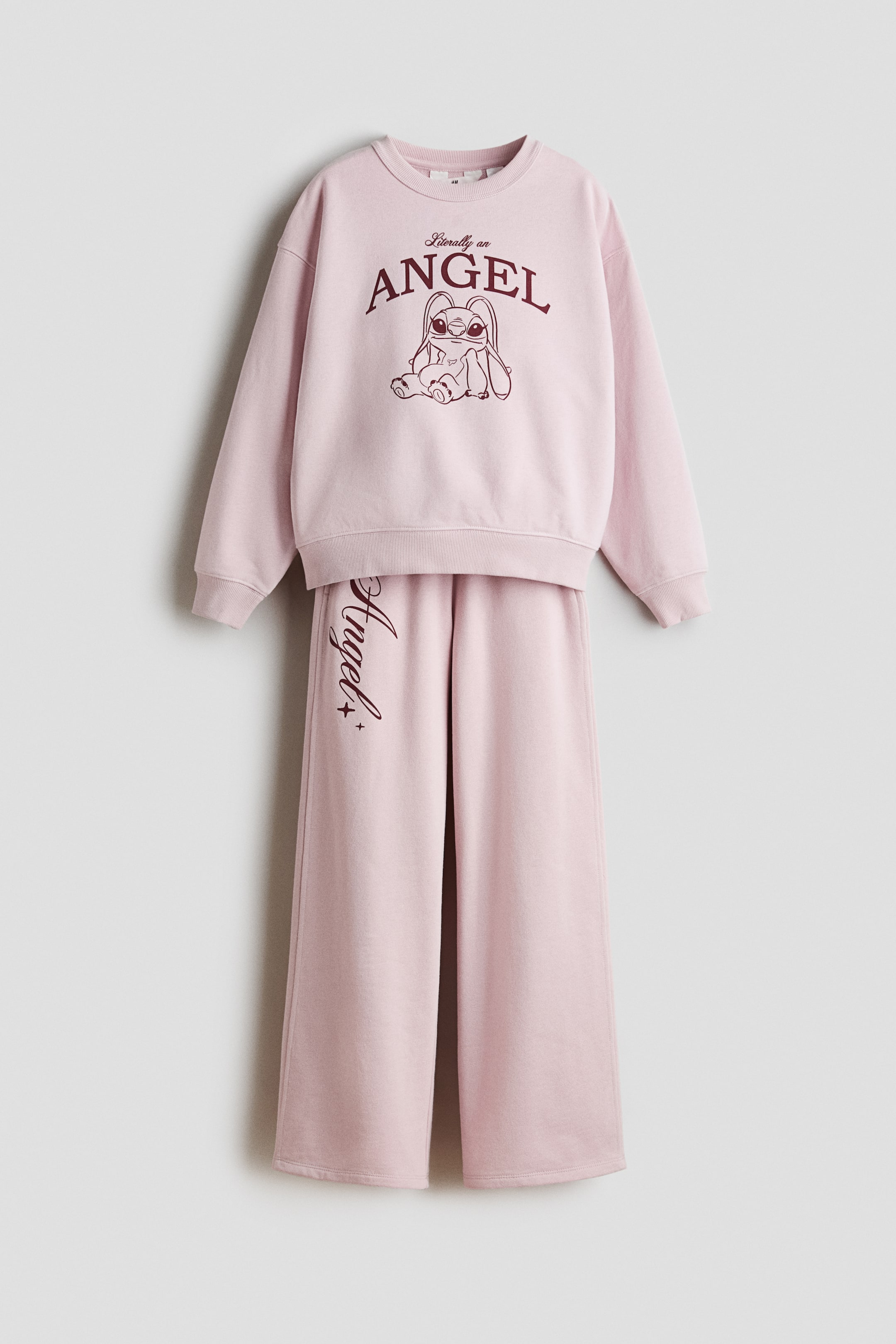 Voir une image plus grande: Un survêtement rose est présenté de face, comprenant un sweat-shirt à col rond et un pantalon à jambes larges. Le sweat-shirt présente des poignets côtelés et un ourlet côtelé, et est orné d'un graphique rouge foncé du personnage Angel et du texte "Littéralement un ANGE." Le pantalon assorti à jambes larges présente un ourlet droit et "Angel" écrit verticalement en rouge foncé sur la jambe gauche.
