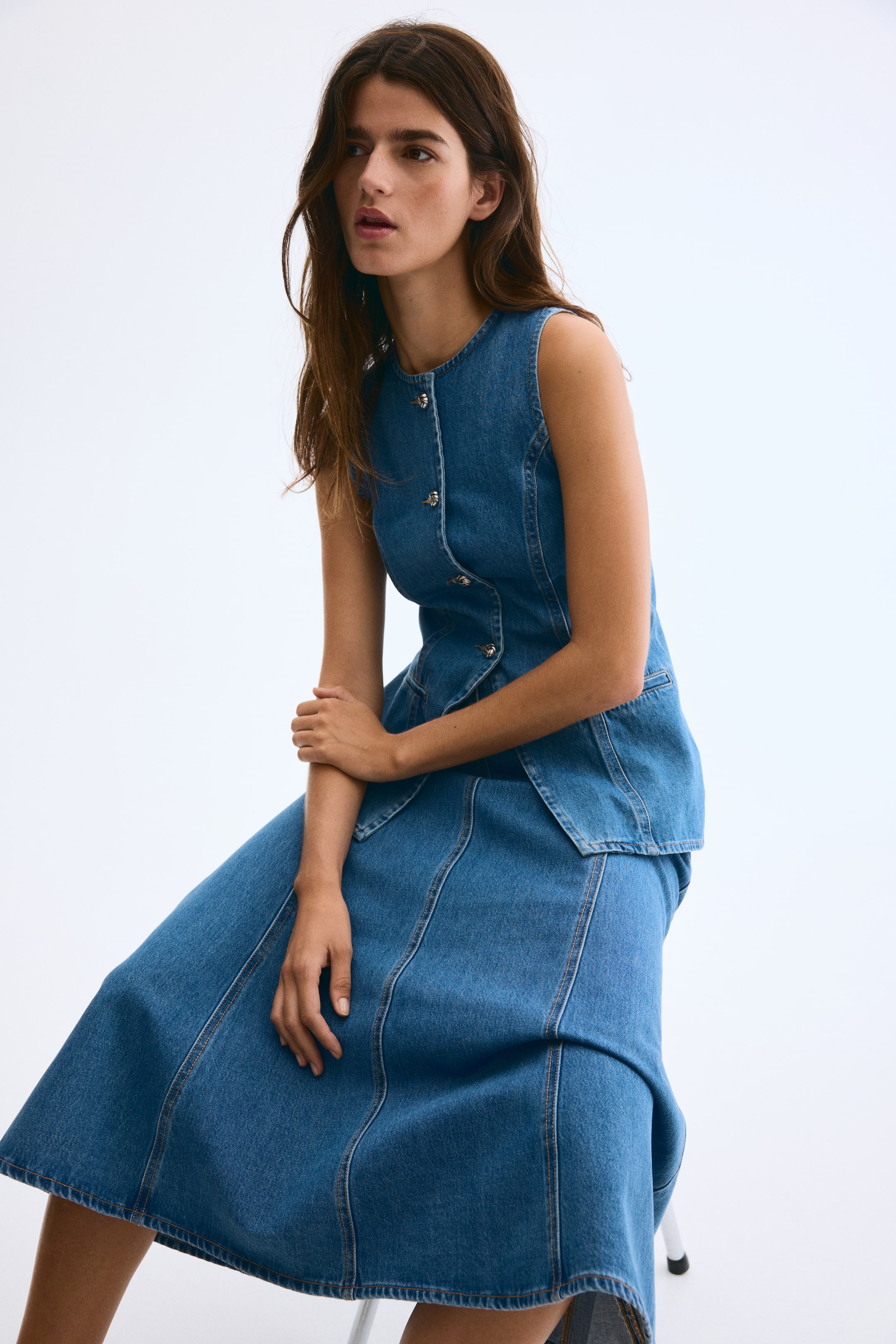 View larger image: A denim dress is being presented from a front, slightly angled view. It features a sleeveless top with a round neckline, silver-toned buttons down the front, and a fitted bodice that flares into a peplum-like overlay at the waist. The midi-length skirt extends below the knees, showcasing an A-line silhouette with vertical seam detailing.