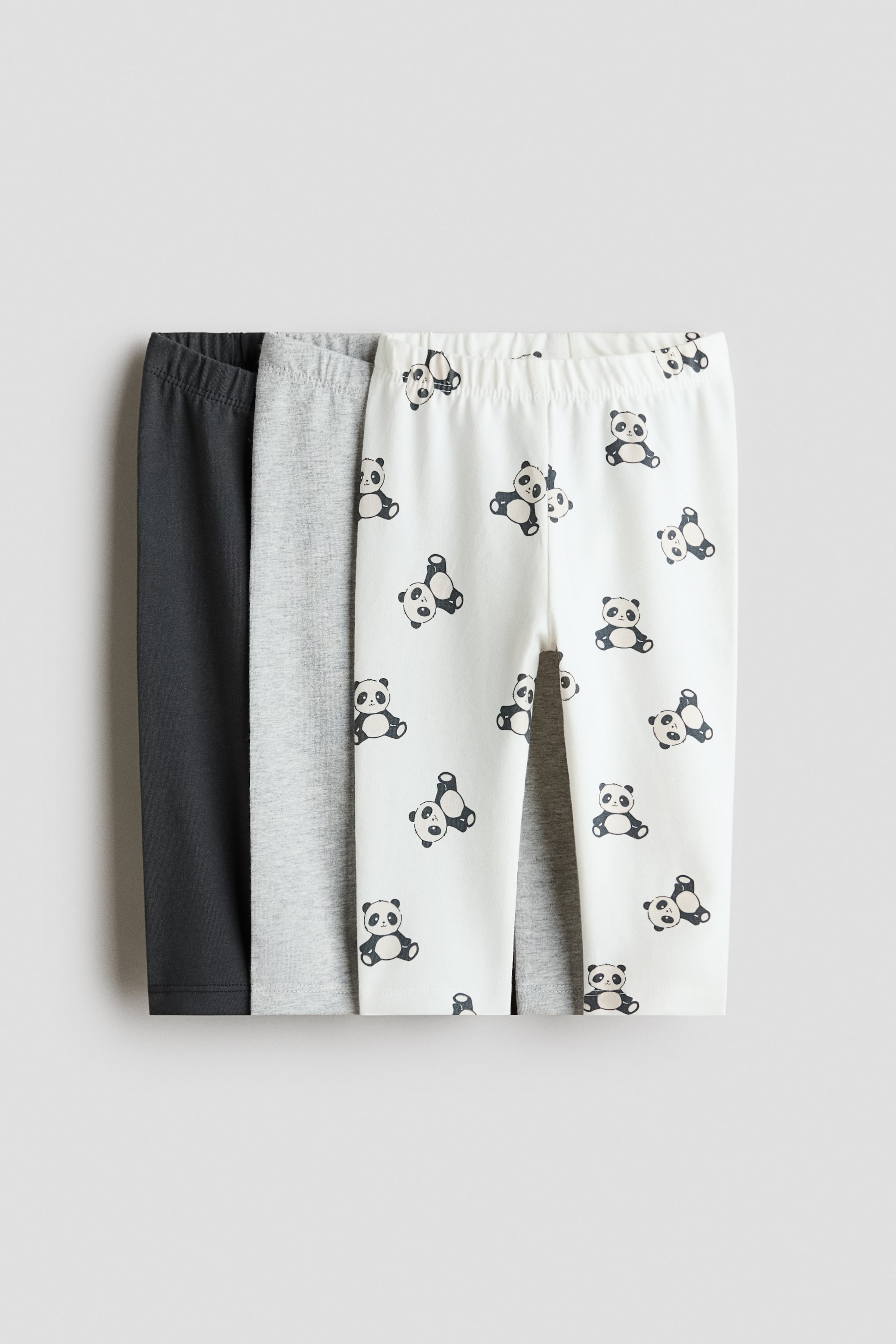 Grotere afbeelding bekijken: Leggings in zachte katoenen jersey zijn van voren afgebeeld in een gelaagde set van drie. Het voorste paar is gebroken wit en voorzien van een all-over print met panda-illustraties, terwijl het middelste paar lichtgrijs gemêleerd is en het achterste paar effen donkergrijs, alle drie met elastische taillebanden.