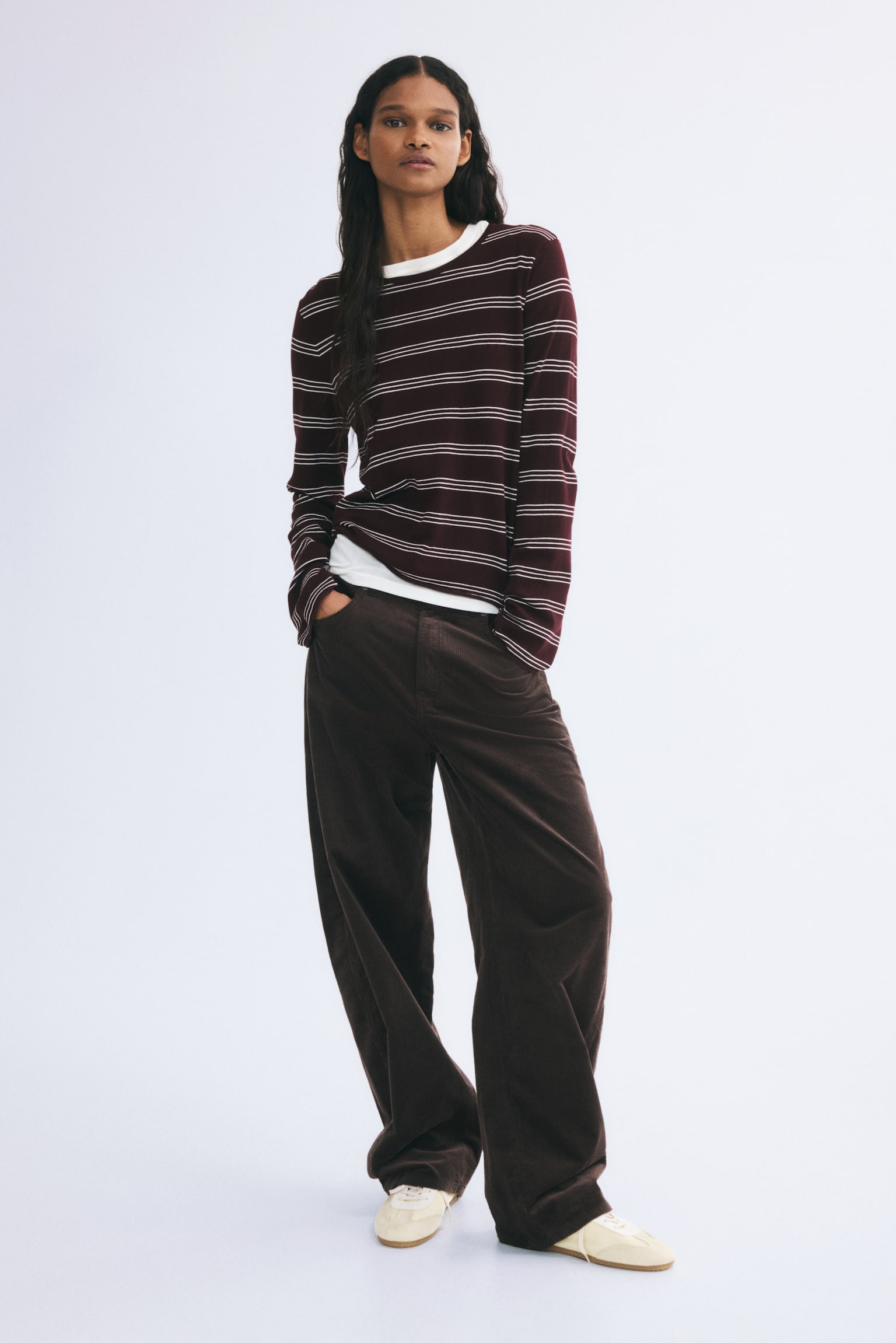 A long-sleeved, fine-knit top in dark red with thin white horizontal stripes is worn by the model. The top features a round neckline and rolled edges, layered over a white crew-neck T-shirt and paired with wide-leg brown corduroy trousers. - 1