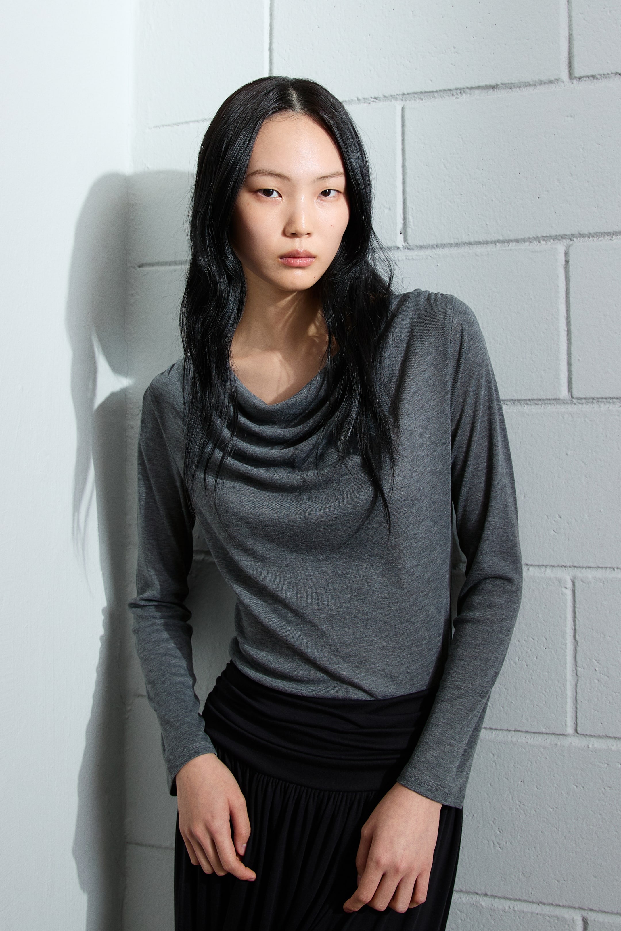 이미지 확대 보기: 어두운 회색의 긴 소매 상의는 드레이프 카울 넥과 몸에 딱 맞는 핏을 특징으로 하며, 블랙 스커트에 넣어 스타일링되었습니다.