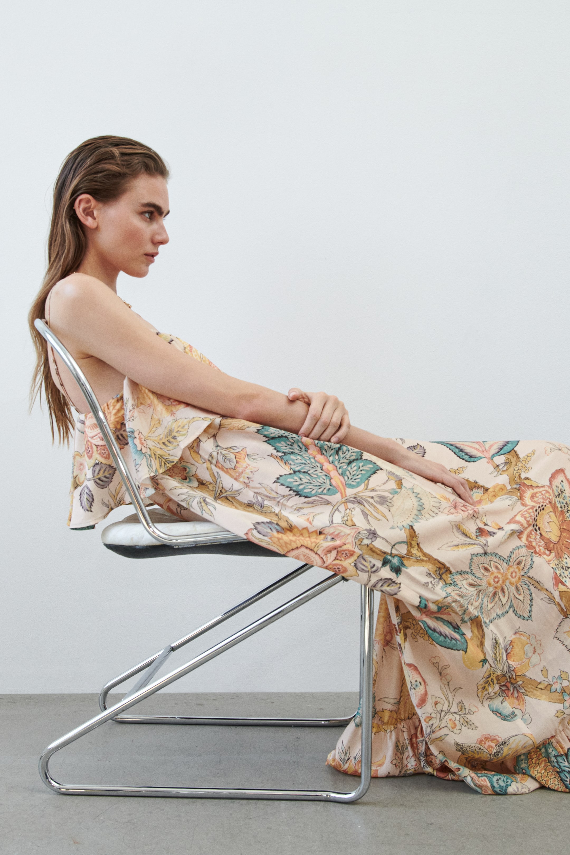 View larger image: The maxi dress features a light peach fabric adorned with an intricate floral and botanical pattern in teal, orange, and yellow hues. It showcases delicate spaghetti straps, a relaxed flounce detail at the bodice, and a flowing tiered skirt that drapes over a chair, revealing its full length and volume.