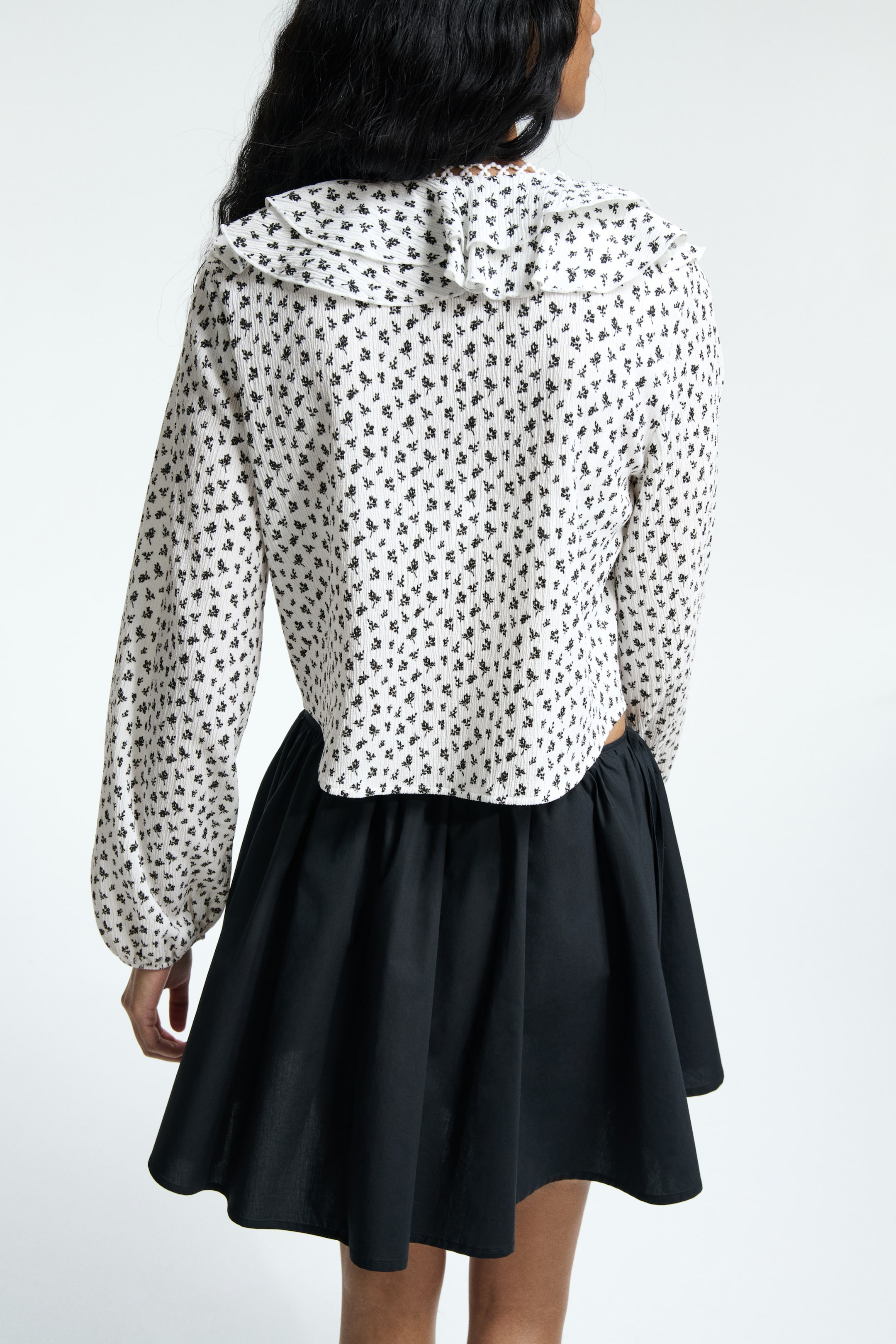 View larger image: A white, ribbed top is showcasing a small black floral pattern and a wide ruffled collar, with its back facing the viewer. It is featuring long sleeves and a cropped hem, with a black, gathered mini skirt being worn below it.