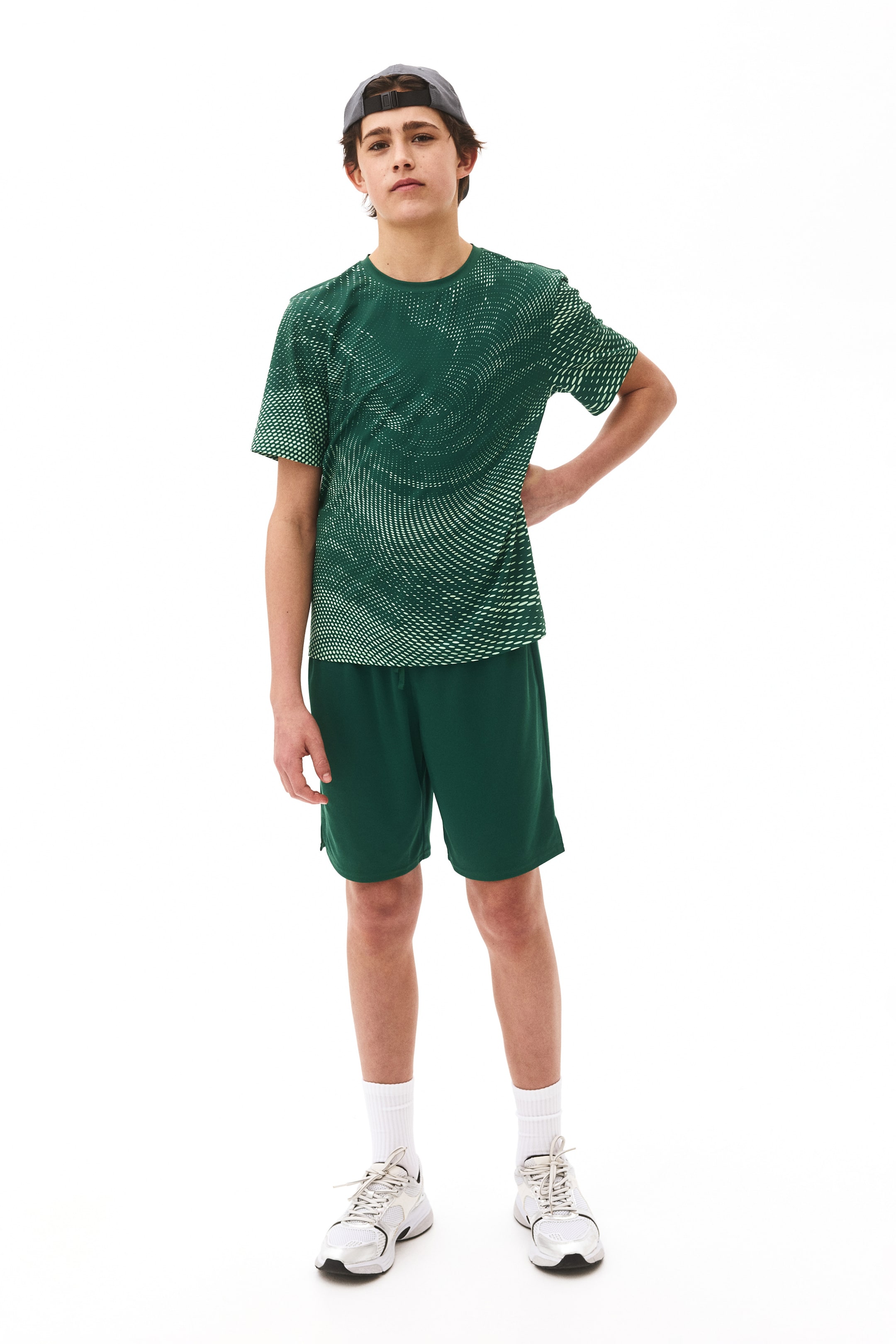 View larger image: A complete athletic ensemble is showcased facing forward, featuring a dark green short-sleeved crew-neck t-shirt adorned with a light green geometric dot pattern forming concentric swirls, paired with solid dark green athletic shorts. A grey baseball cap is worn backward, complemented by white crew socks and white and grey athletic shoes.