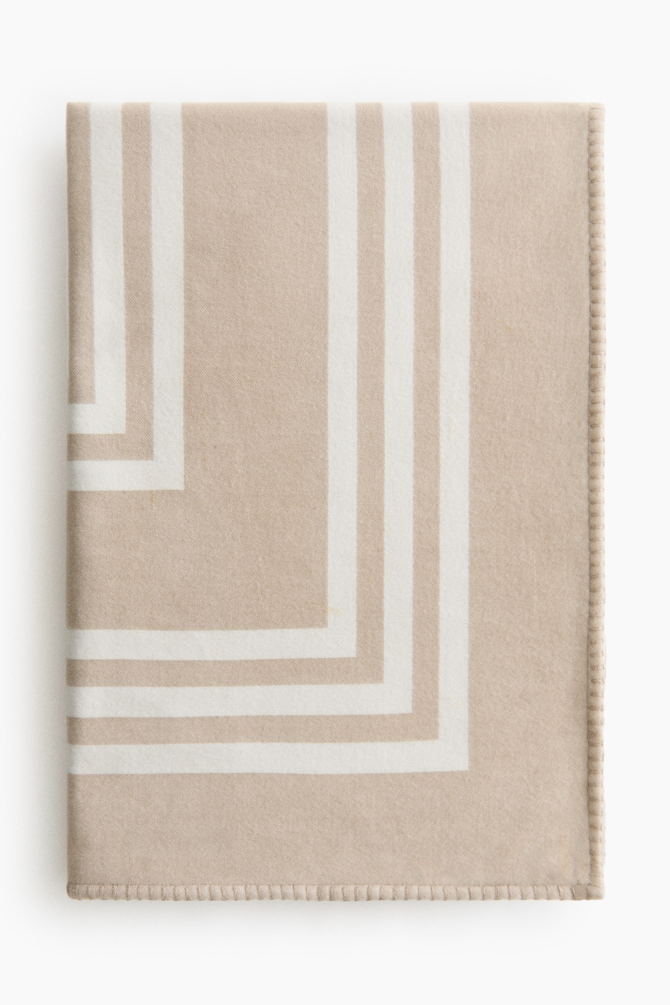 拡大画像を表示: ブランケットはたたまれて置かれ、柔らかなベージュの編み目が白いグラフィックデザインの層状の長方形のストライプを特徴としている。また、ブランケットの縁にはブランケットステッチが施されている。