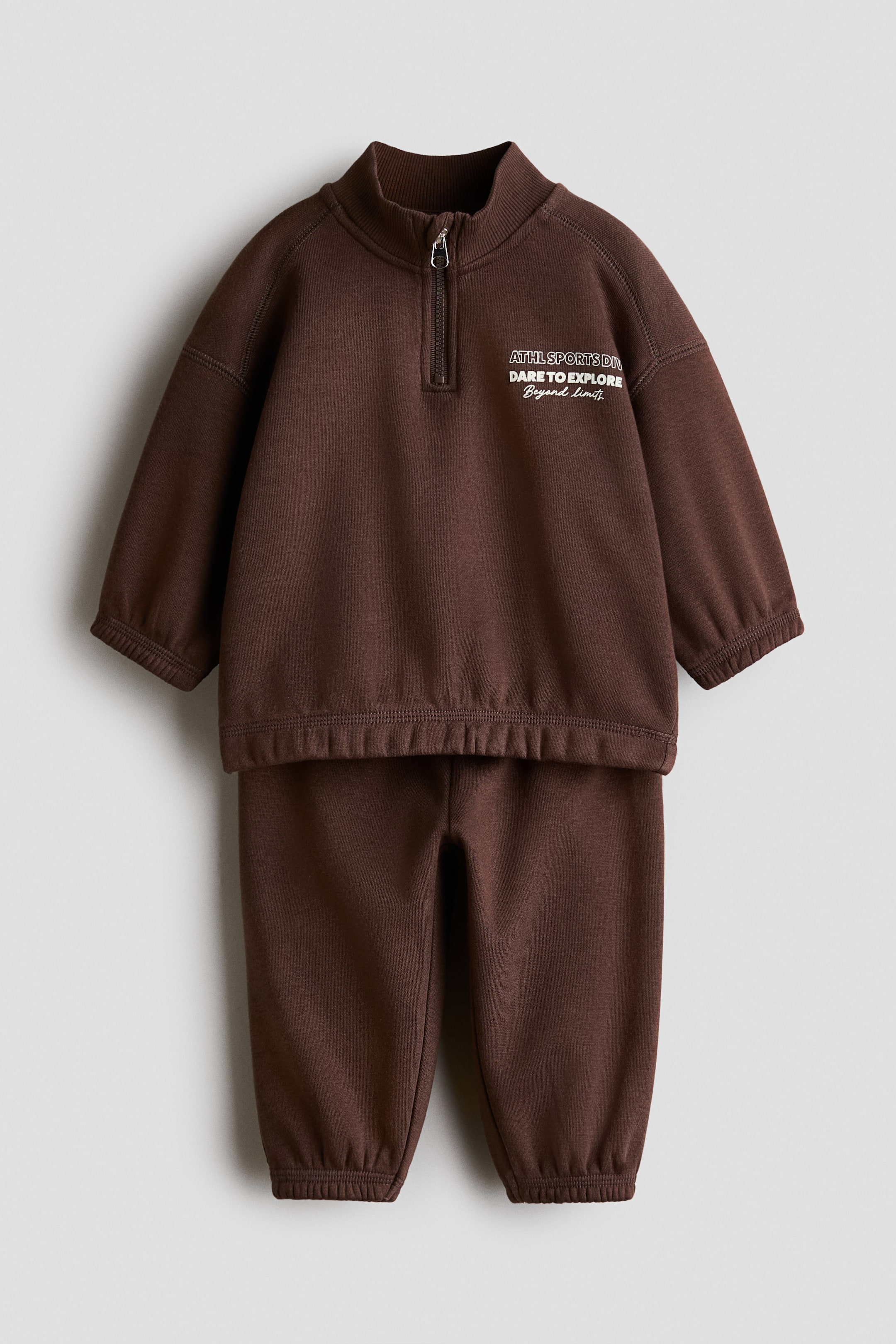 Voir une image plus grande: Un ensemble de sweatshirt en molleton brun comprend un haut avec un petit imprimé blanc sur le devant indiquant « ATHL SPORTS DIV DARE TO EXPLORE Beyond limits », un col montant côtelé avec demi-fermeture éclair, des manches longues raglan et une bande élastique recouverte aux poignets et à l’ourlet. Le jogging assorti présente une bande élastique recouverte aux bas de jambe.