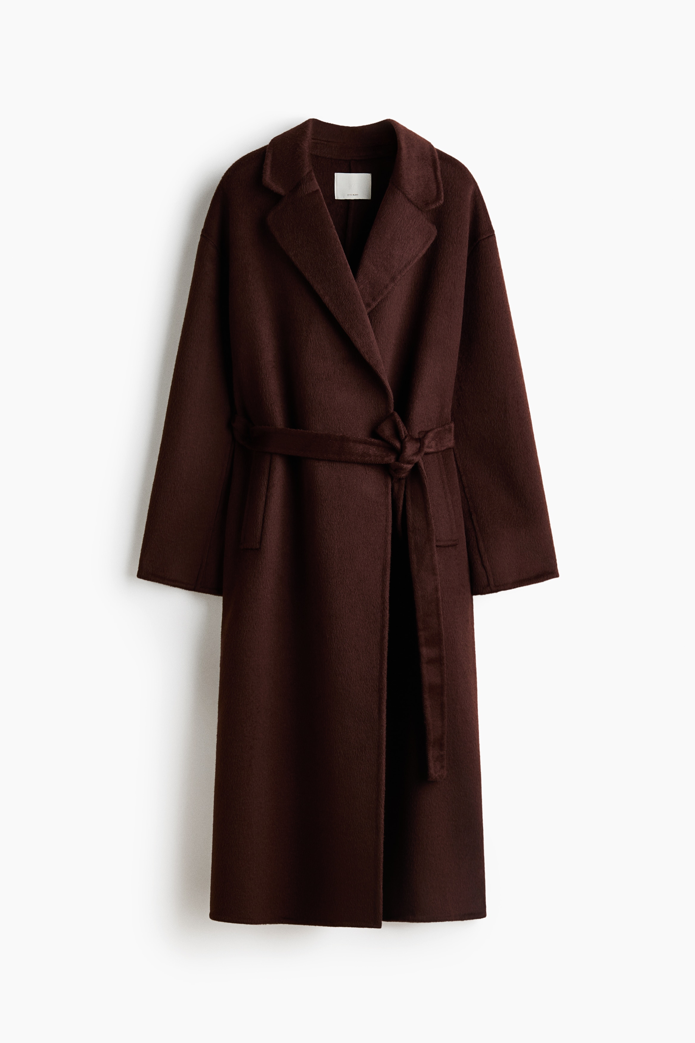 A long, dark brown wool-blend coat featuring an oversized fit, notch lapels, and a tied waist belt is worn by the model. The coat is layered over a light-coloured collared shirt and dark burgundy leather trousers, with a matching dark leather shoulder bag worn over one shoulder.