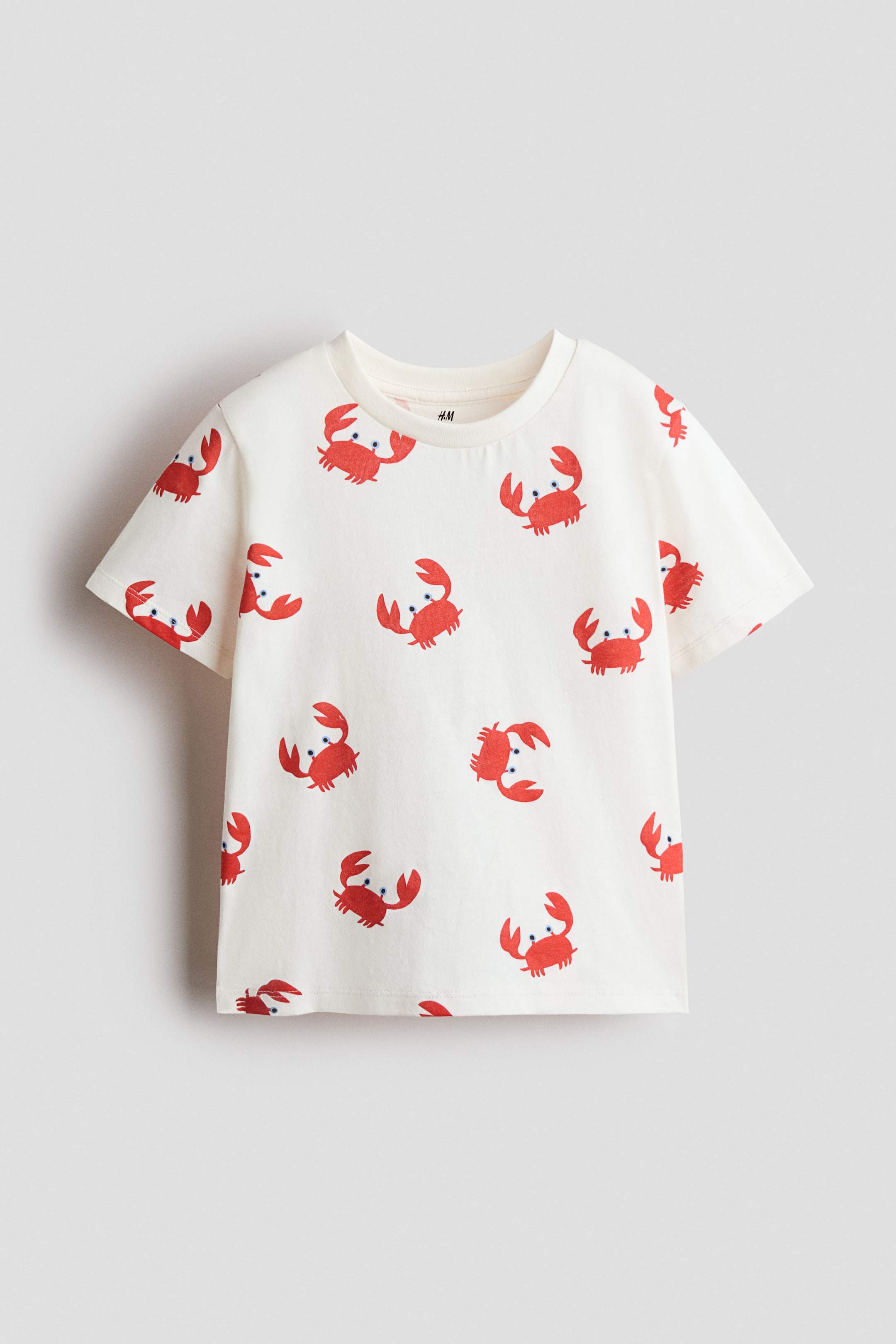 拡大画像を表示: Tシャツは、青い目を持つ赤いカニの総柄が全面に描かれたデザインで、正面からのビューで表示されています。柔らかいコットンジャージー素材で作られており、ネックライン周りにはリブが施され、ドロップショルダーとストレートカットの裾が特徴です。