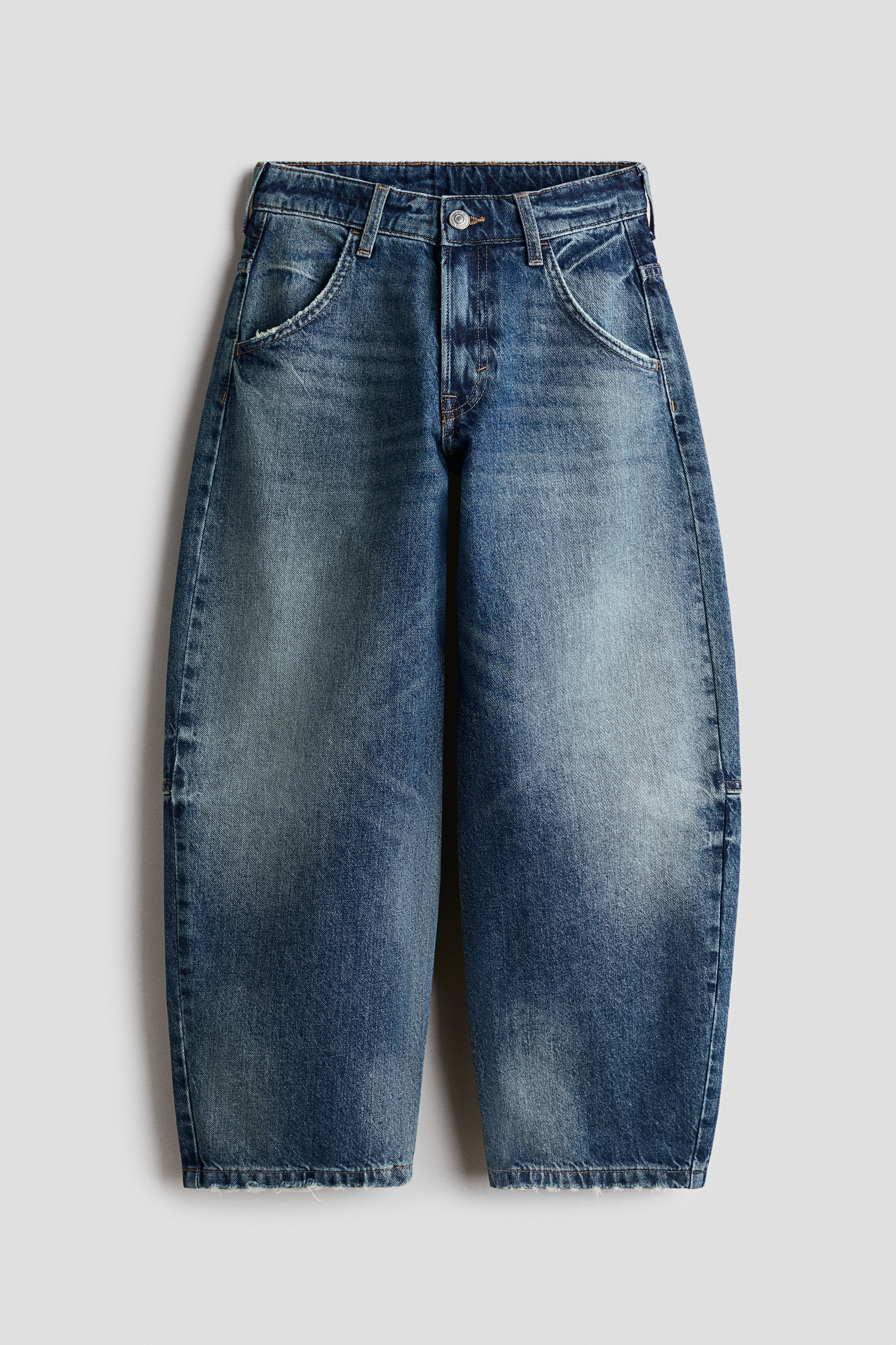 Jeans in washed blue cotton denim are shown from the front, featuring a loose fit through the hips and thighs with curved barrel legs. The jeans are displaying a faded finish on the thighs and are styled with a grey hoodie partially covering the waistband.