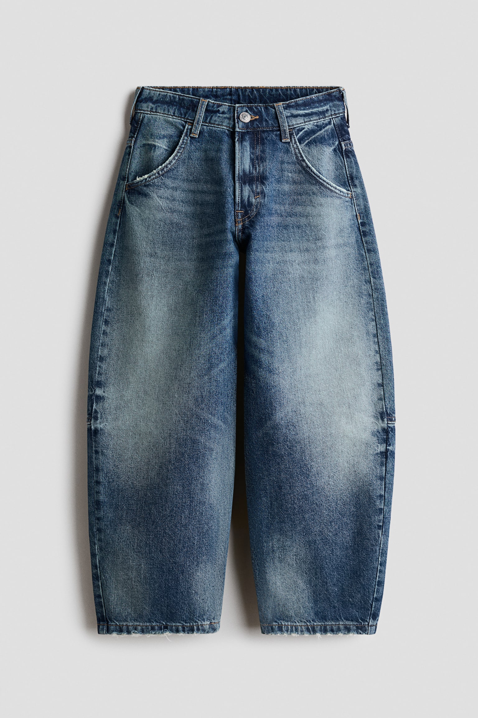 A pair of blue denim jeans is shown from the front, showcasing a balloon-leg silhouette with a faded, worn-in wash on the thighs and knees. The jeans feature classic five-pocket styling, contrast stitching, a button fastening with a zip fly, and slightly frayed hems.