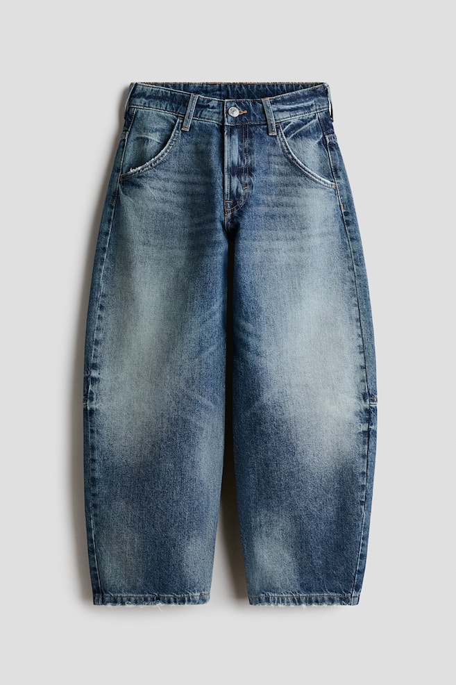 A pair of blue denim jeans is shown from the front, showcasing a balloon-leg silhouette with a faded, worn-in wash on the thighs and knees. The jeans feature classic five-pocket styling, contrast stitching, a button fastening with a zip fly, and slightly frayed hems. - 1