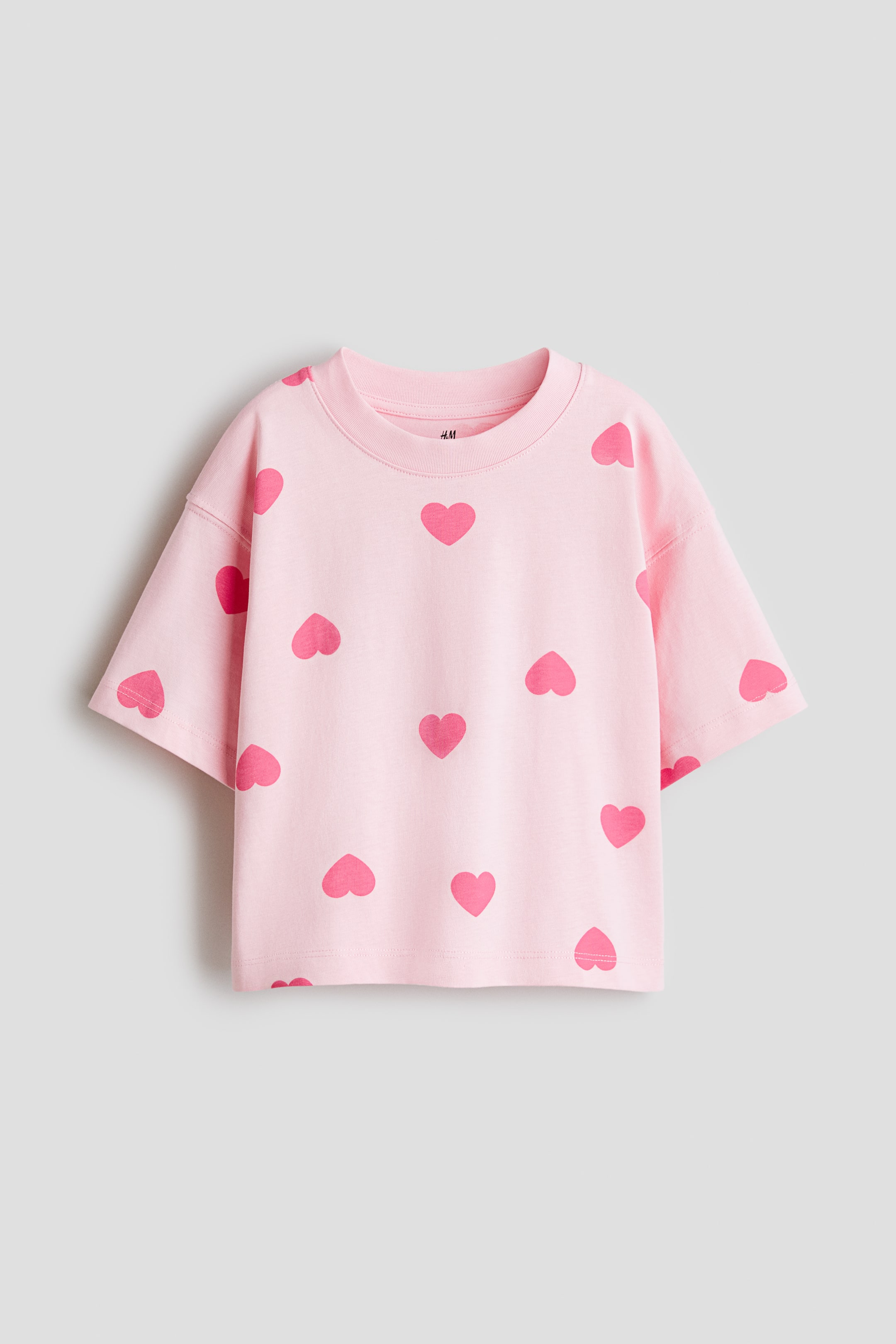 View larger image: A light pink t-shirt, FRONT, is featuring a ribbed crew neck and short, dropped-shoulder sleeves. The t-shirt is patterned all over with darker pink hearts.
