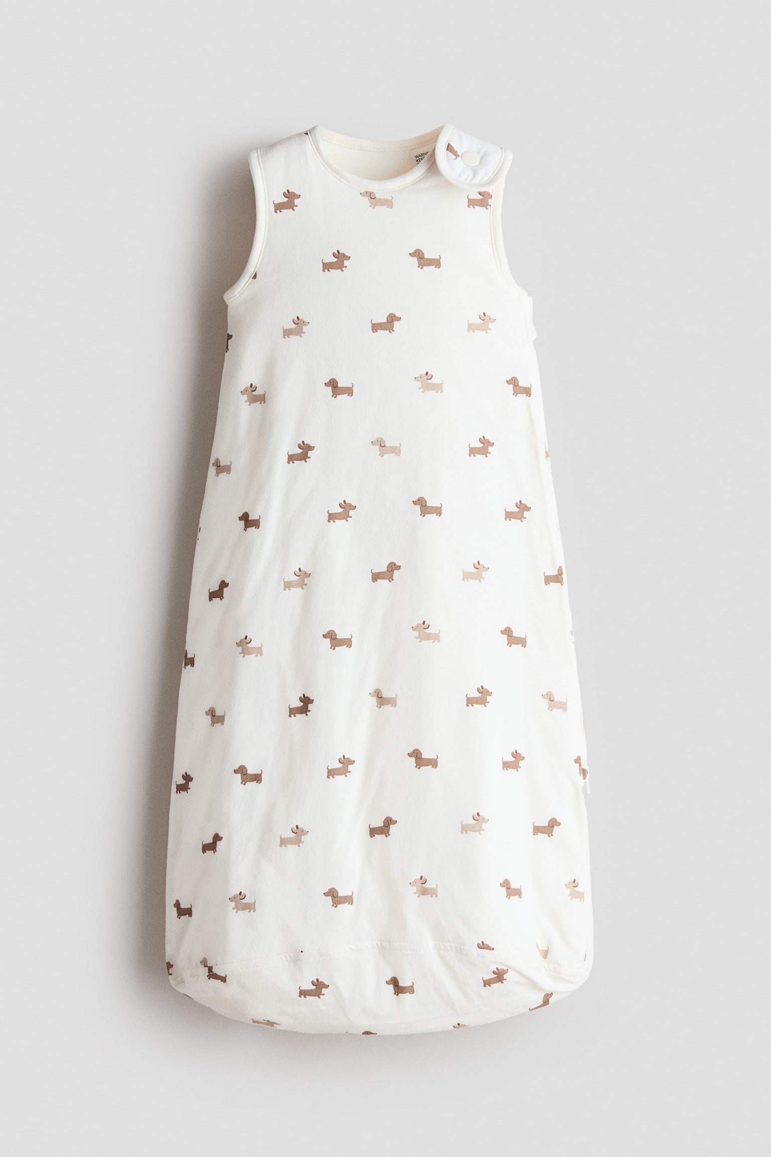 A padded sleep bag in cream cotton jersey featuring an all-over print of small brown dachshunds in a front-facing view. The sleeveless garment features a rounded neckline, a press-stud fastening on one shoulder, and a side zip.
