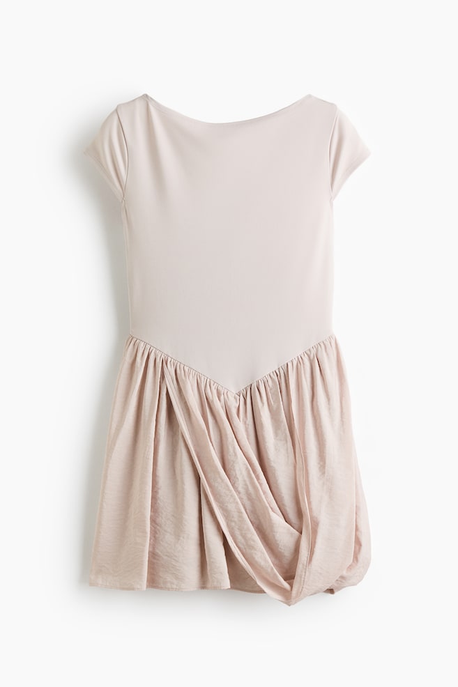 Ein blassrosa Kleid präsentiert ein glattes, mattes Oberteil mit Bootsausschnitt und kurzen Ärmeln. Dieses Oberteil ist durch eine V-förmige Naht mit einem gerafften und drapierten Rockteil verbunden, der einen glänzenderen Stoff mit voluminösen Falten zeigt. - 1