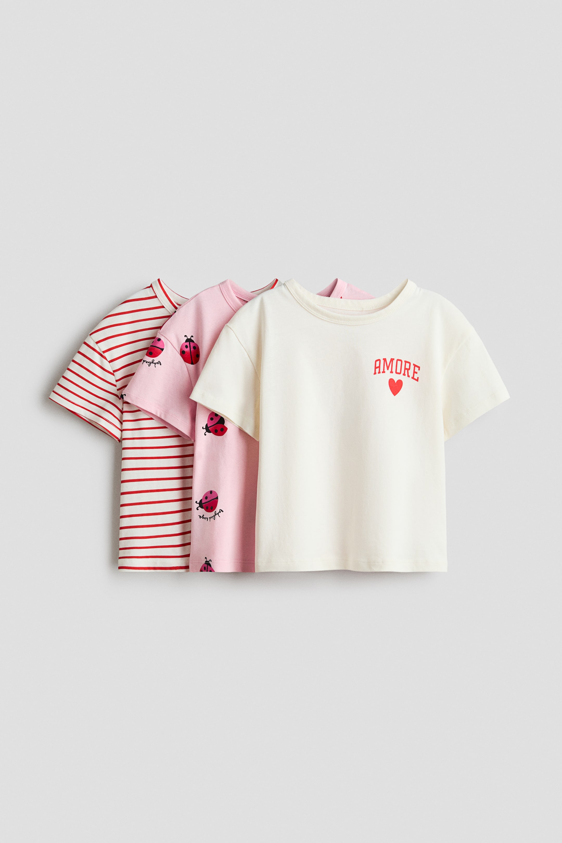 View larger image: Three short-sleeved t-shirts are displayed in a layered arrangement, angled slightly to the right. The front t-shirt is cream, featuring red "AMORE" text and a heart; the middle t-shirt is light pink, showing scattered red ladybug graphics; and the back t-shirt is white, displaying red horizontal stripes.