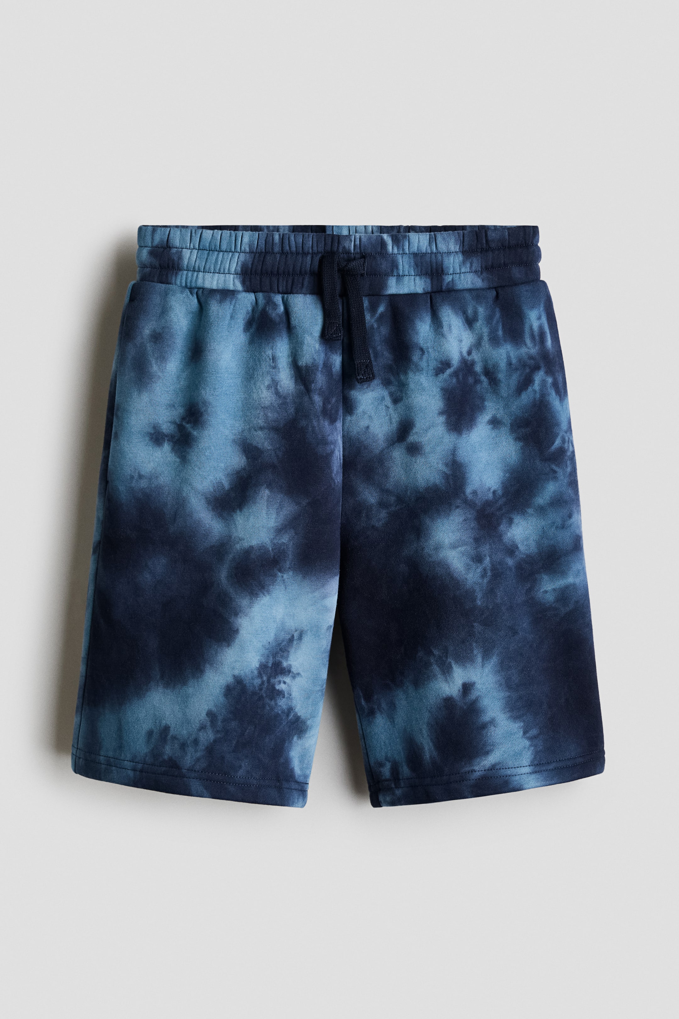 View larger image: The shorts feature a blue tie-dye pattern with varying shades of light and dark blue. They are designed with an elasticated waistband, including a visible dark blue drawstring, and straight legs ending in stitched hems.