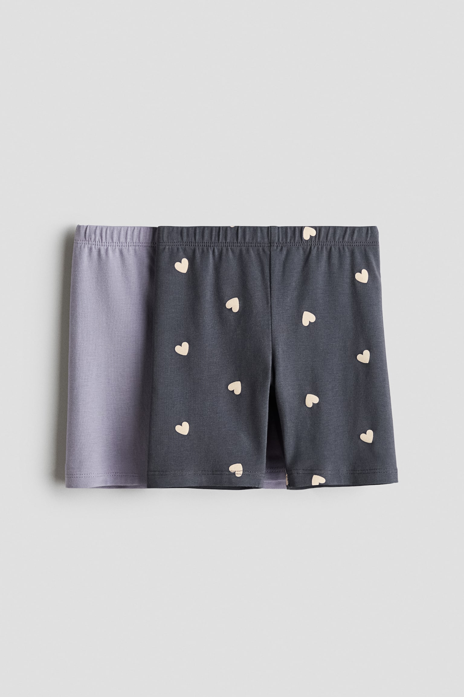 Hai chiáº¿c quáº§n short xe Äáº¡p báº±ng cotton jersey ÄÆ°á»£c trÆ°ng bÃ y á» gÃ³c nhÃ¬n chÃ­nh diá»n, vá»i má»t chiáº¿c mÃ u tÃ­m nháº¡t trÆ¡n ÄÆ°á»£c xáº¿p lá»p phÃ­a sau má»t chiáº¿c mÃ u xÃ¡m Äáº­m cÃ³ há»a tiáº¿t trÃ¡i tim mÃ u kem nhá» phá»§ toÃ n bá» bá» máº·t. Cáº£ hai chiáº¿c Äá»u cÃ³ cáº¡p thun co giÃ£n vÃ  gáº¥u may tháº³ng.