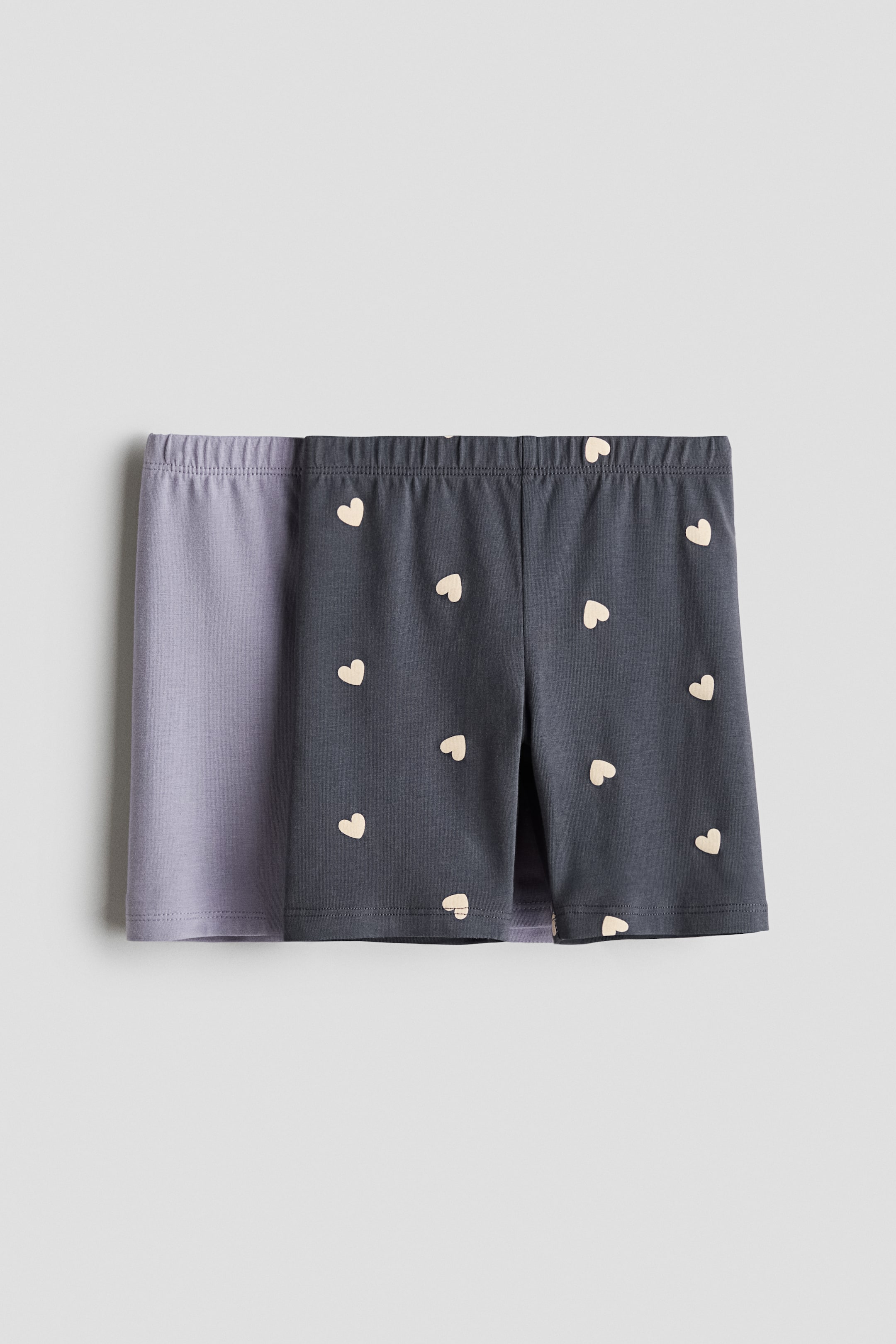 Xem ảnh lớn hơn: Hai chiếc quần short xe đạp bằng cotton jersey được trưng bày ở góc nhìn chính diện, với một chiếc màu tím nhạt trơn được xếp lớp phía sau một chiếc màu xám đậm có họa tiết trái tim màu kem nhỏ phủ toàn bộ bề mặt. Cả hai chiếc đều có cạp thun co giãn và gấu may thẳng.
