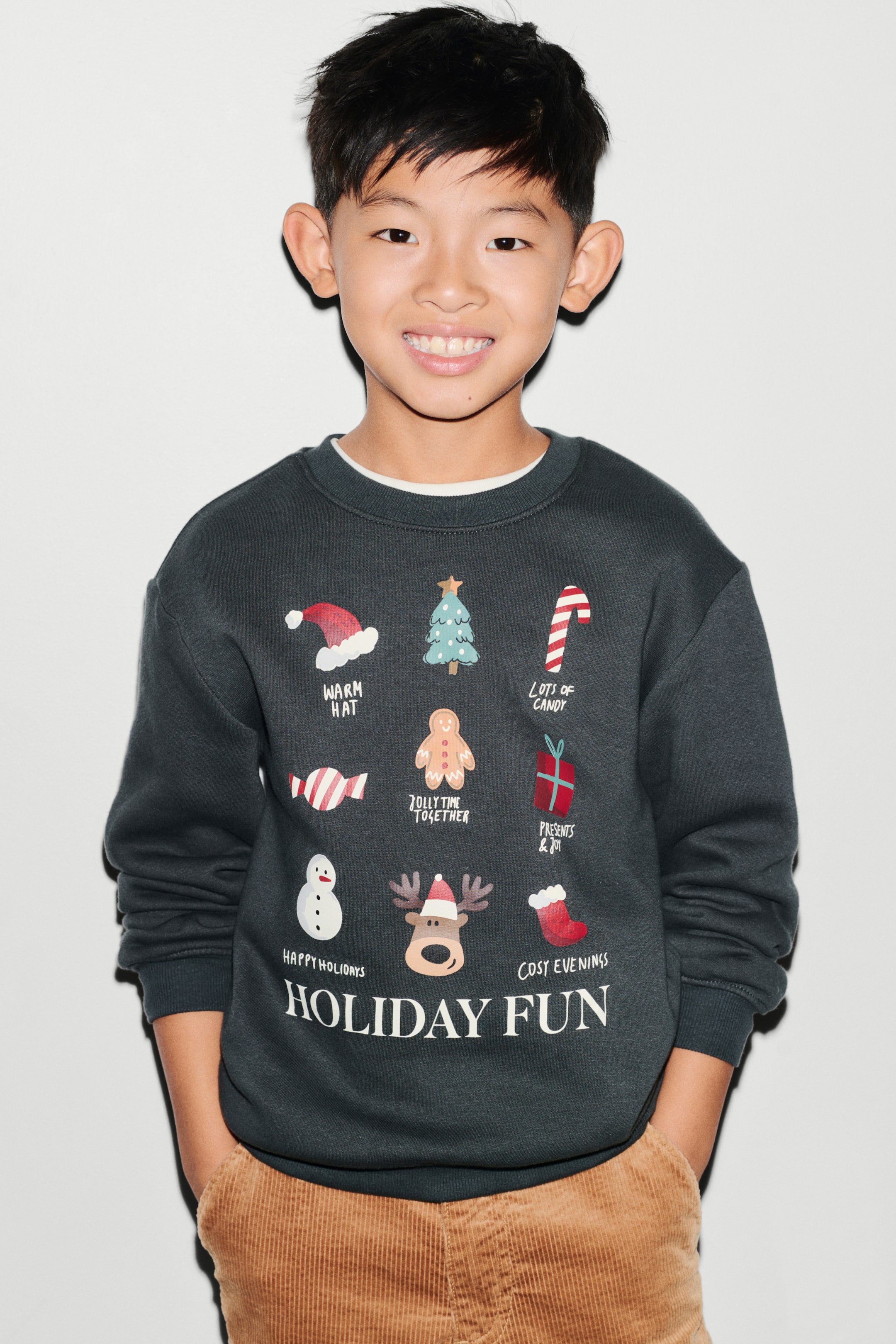View larger image: A dark grey sweatshirt is shown from the front, featuring a round, rib-trimmed neckline, dropped shoulders, and long sleeves with ribbed cuffs. The chest displays a printed holiday-themed design with multiple festive illustrations and the text "HOLIDAY FUN" at the bottom.
