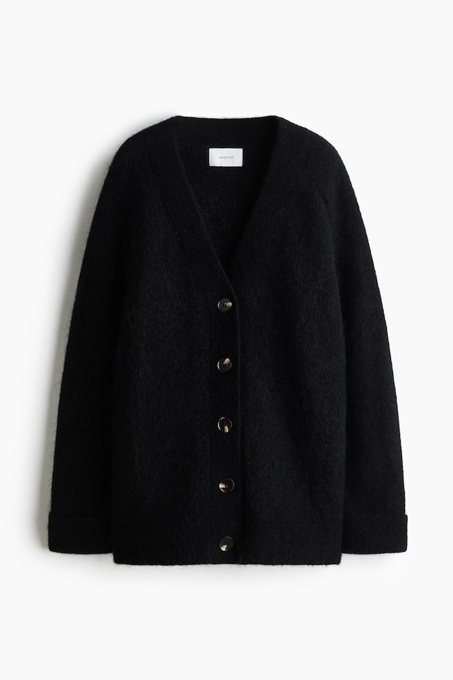 A black oversized cardigan in a fuzzy knit is facing front, featuring a deep V-neckline and five dark, marbled buttons down the front placket. The cardigan shows long raglan sleeves, ribbed cuffs, and a ribbed hem. - 1