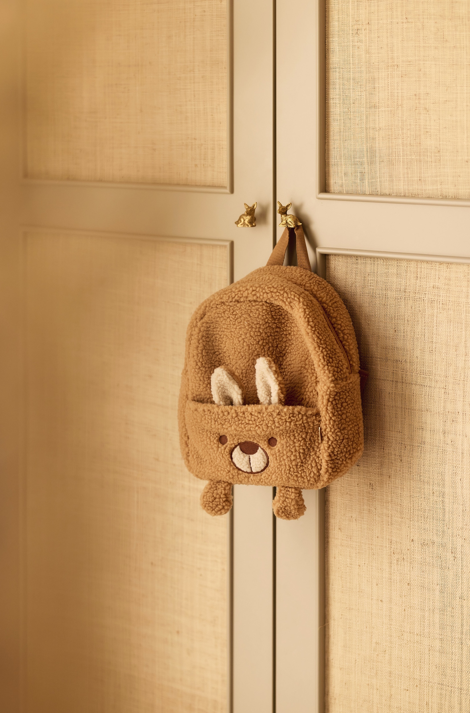Een van voren gefotografeerde kinder-rugzak met een bruine sherpa fleece buitenkant, vormgegeven als een beer. Het voorvak met rits toont een geborduurd gezicht met zachte rechtopstaande oren en kleine pootjes aan de onderkant, terwijl een beige webbing lus als handgreep aan de bovenkant dient.