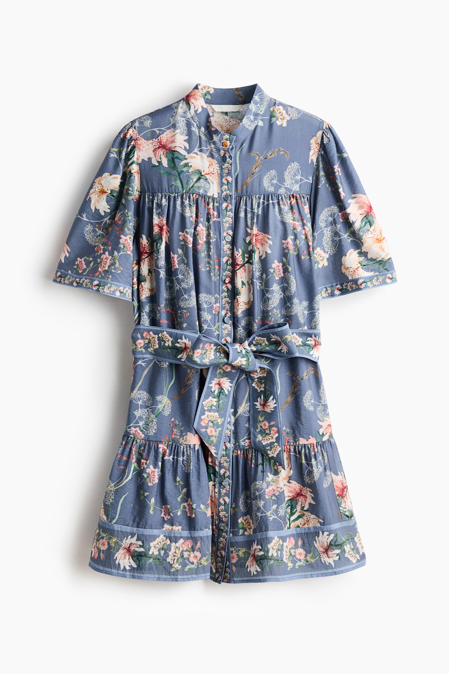 O rochie este prezentată într-o vedere frontală, afișând un material de bază albastru cu un model complet de crini roz, piersică și albi și un frunziș delicat. Este proiectată cu un guler înalt, o plachetă cu nasturi în față, mâneci scurte bufante cu o bandă cu model și o curea din același material legată la talie, conducând la o fustă cu volane cu o bordură cu model la tiv. - 1