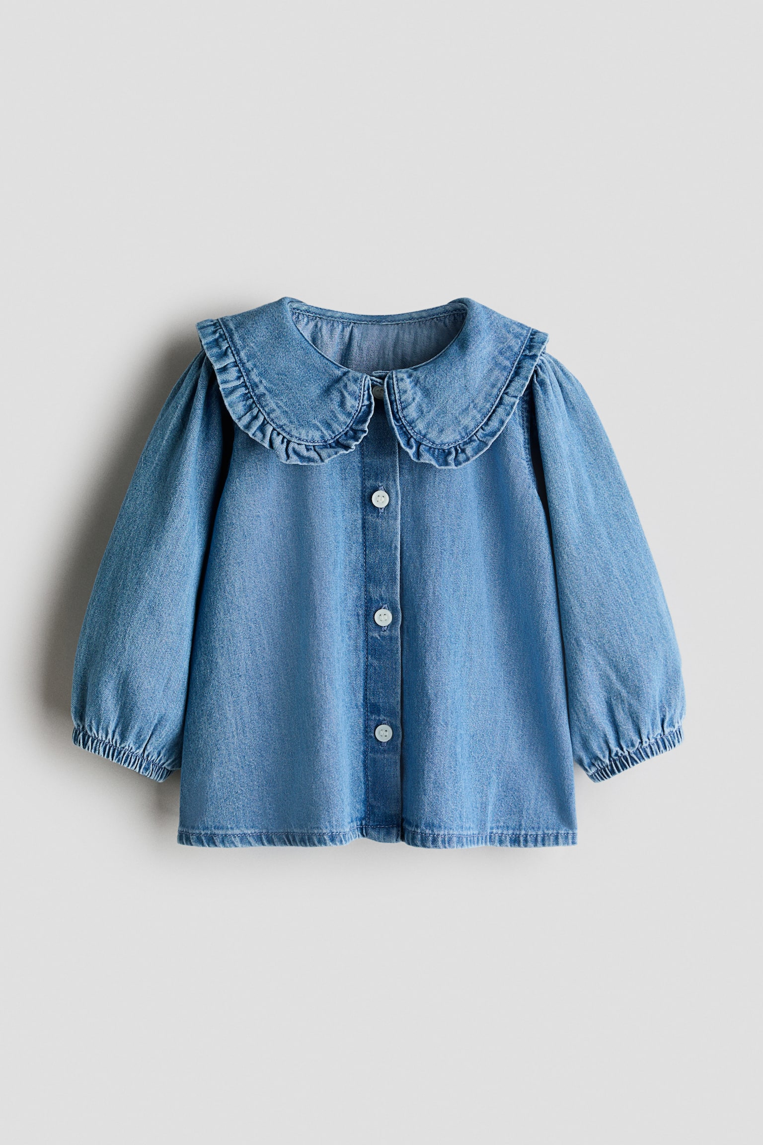 Een lichtblauw denim shirt met een grote, gerimpelde Peter Pan-kraag en lange, gepofte mouwen die zijn verzameld bij de elastische manchetten. Het heeft ook een volledige knoopsluiting met witte knopen aan de voorkant. - 1