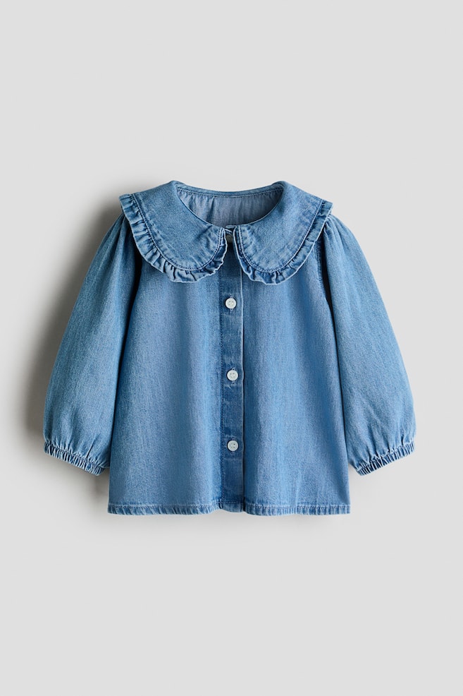 A light blue denim shirt showcases a large, ruffled Peter Pan collar and long, puffed sleeves gathered at the elasticated cuffs. It also features a full button placket with white buttons down the front. - 1
