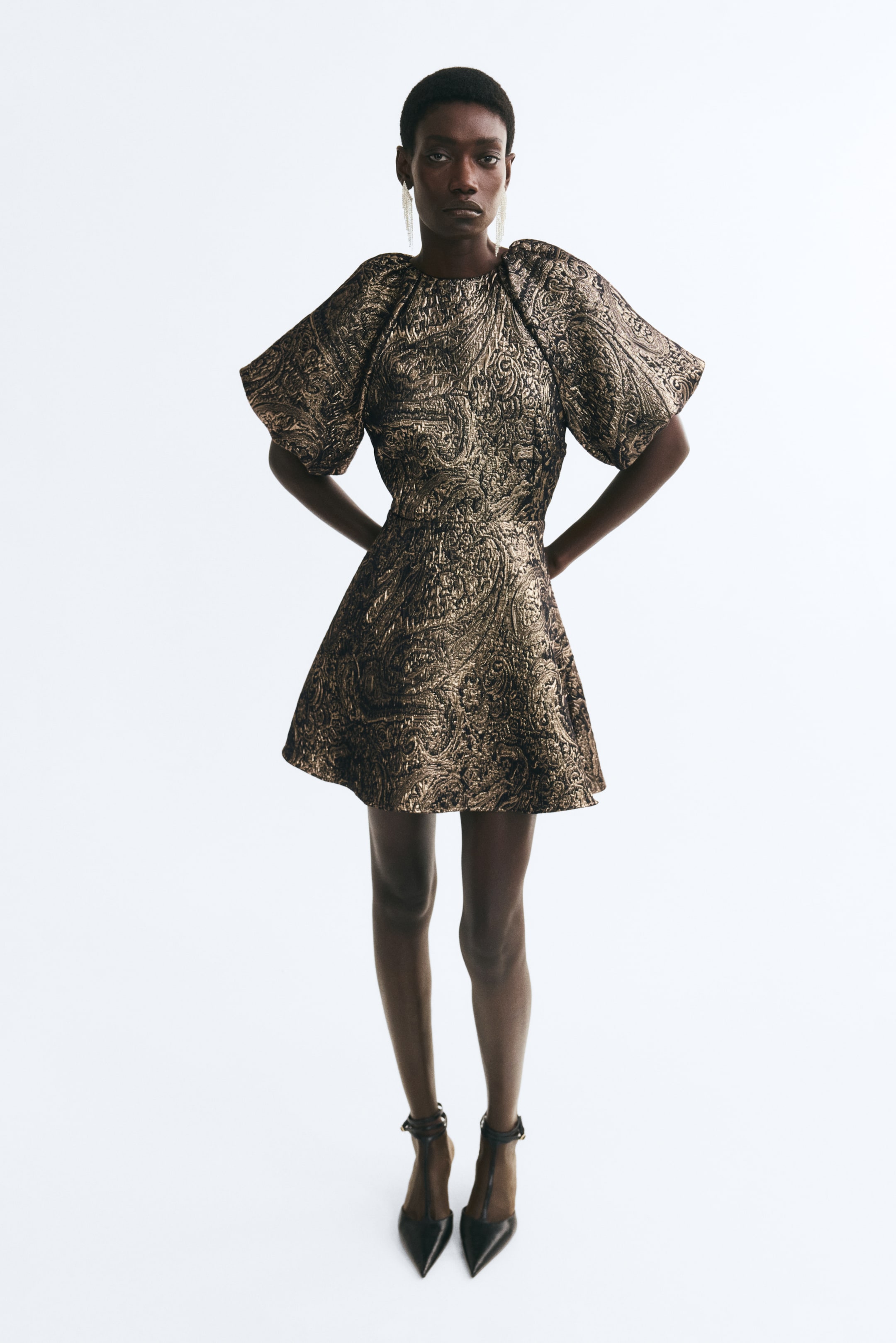 View larger image: A short, fitted dress in a textured gold and black metallic pattern is being worn by the model, who is standing with her hands behind her back. The dress features a round neckline, voluminous short raglan balloon sleeves, and a gently flared skirt.