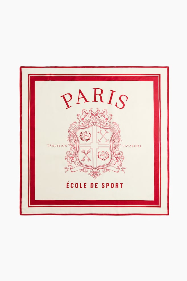 A square scarf in a soft weave is laid flat, showcasing the front design with a cream-coloured base and a red printed pattern. The design features a double red border framing a central crest with equestrian motifs, with the word "PARIS" arched above and "ï¿½COLE DE SPORT" printed below. - 1