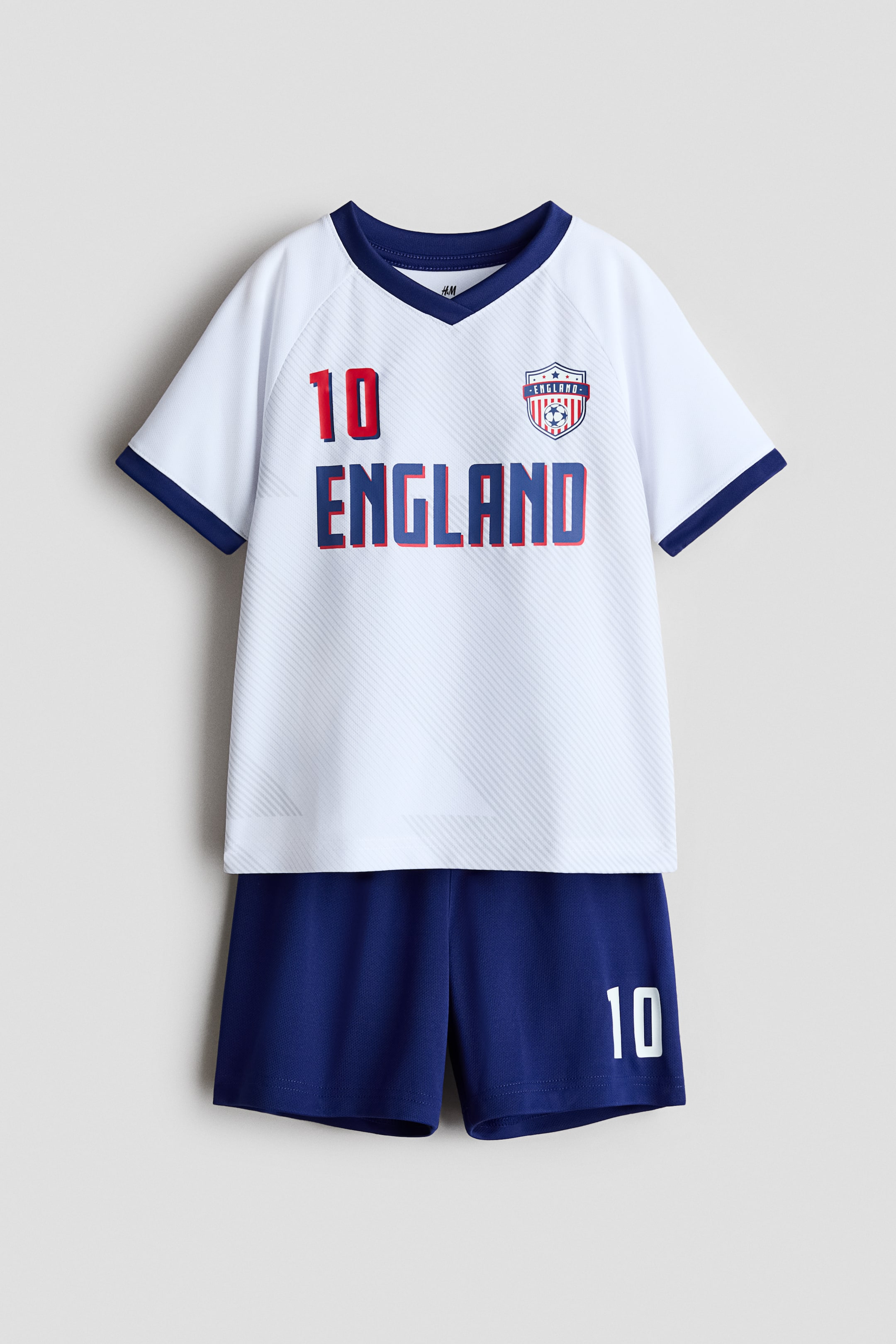 Visa större bild: En FRAMSIDIG vy visar ett tryckt fotbollsställ tillverkat av mjukt mesh-tyg. Den vita kortärmade toppen har en marinblå V-ringning och ärmslut, med texten "10" och "ENGLAND", samt en sköldlogotyp på framsidan, allt över ett subtilt diagonalt texturerat mönster. De matchande marinblå shortsen visar "10" tryckt på nedre högra benet.