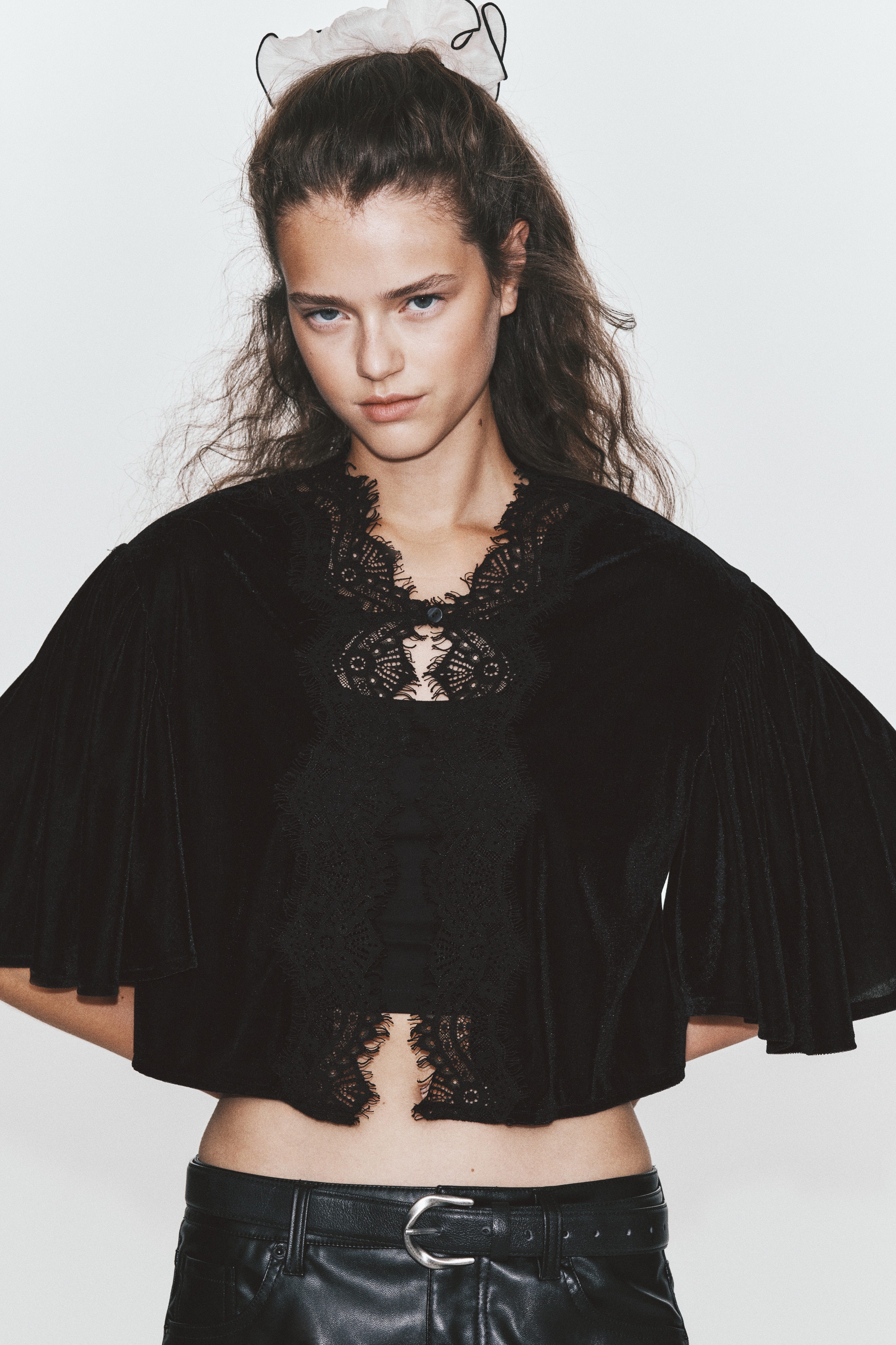 A black velvet cropped top is shown from the front, featuring a high lace-trimmed collar that extends into a decorative lace panel down the centre front. The top has wide, draped short sleeves and a straight cropped hem.