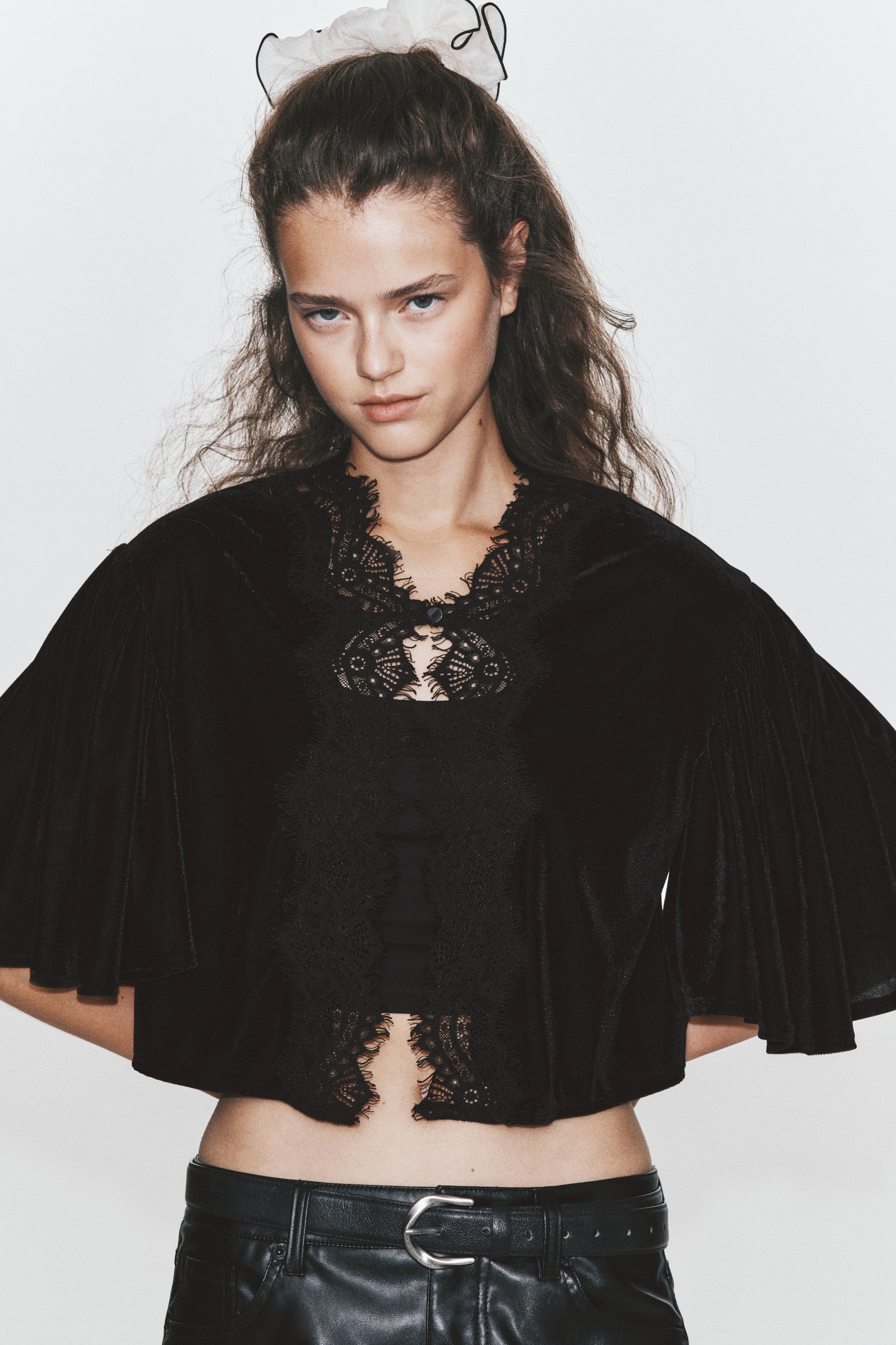 Ampliar la imagen: La modelo lleva una blusa corta de terciopelo negro, con un ribete de encaje con borde ondulado alrededor del escote y a lo largo del frente abierto, el cual se cierra con un solo botón y presilla. La blusa tiene mangas cortas y amplias, y se estiliza sobre una top negra, combinada con pantalones negros con acabado tipo piel.