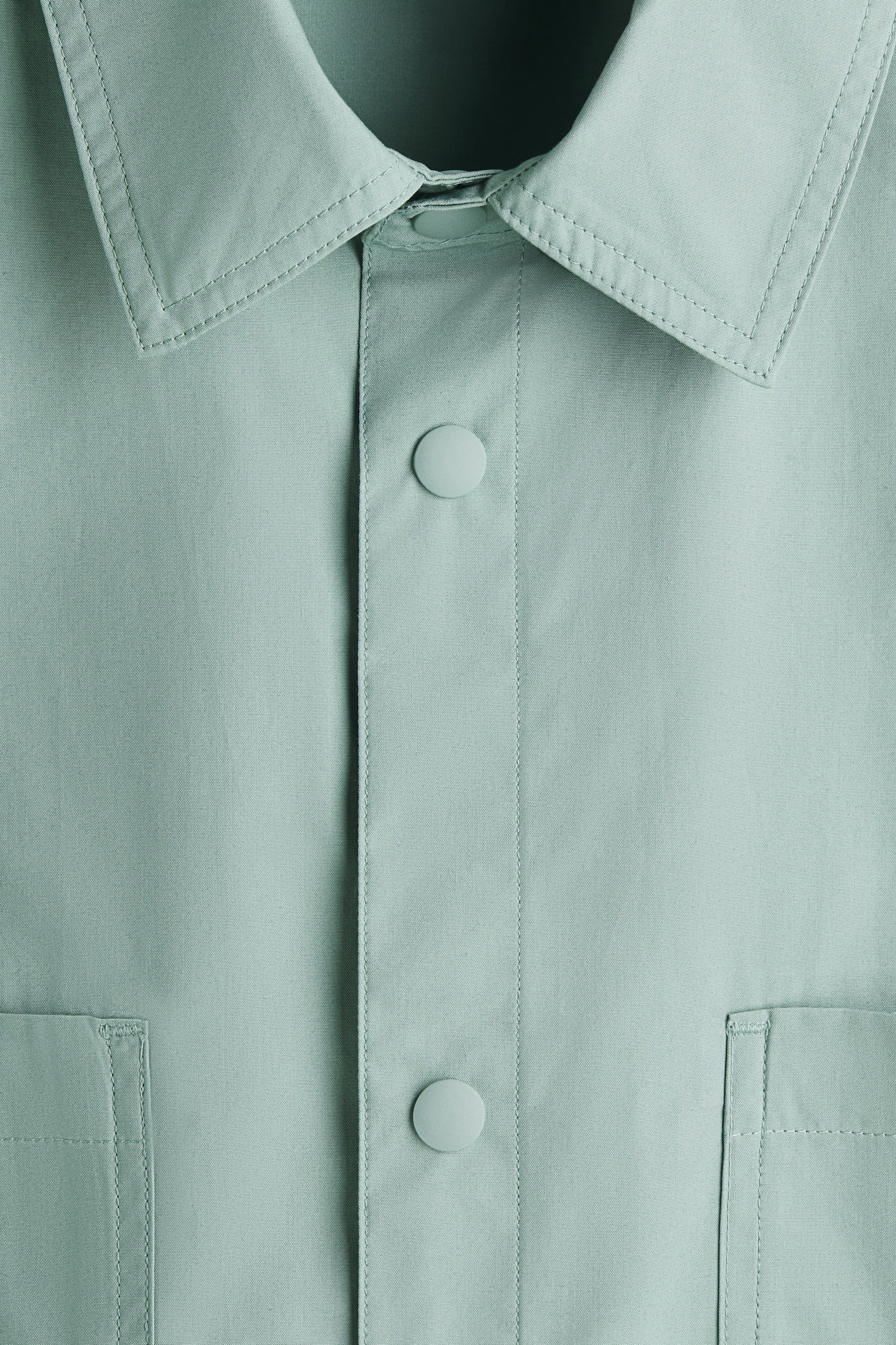 View larger image: A light green shirt is displaying a pointed collar with double-stitched edges and a central placket with two round, light green snap buttons. Two rectangular patch pockets are appearing on the lower front, each featuring visible topstitching.