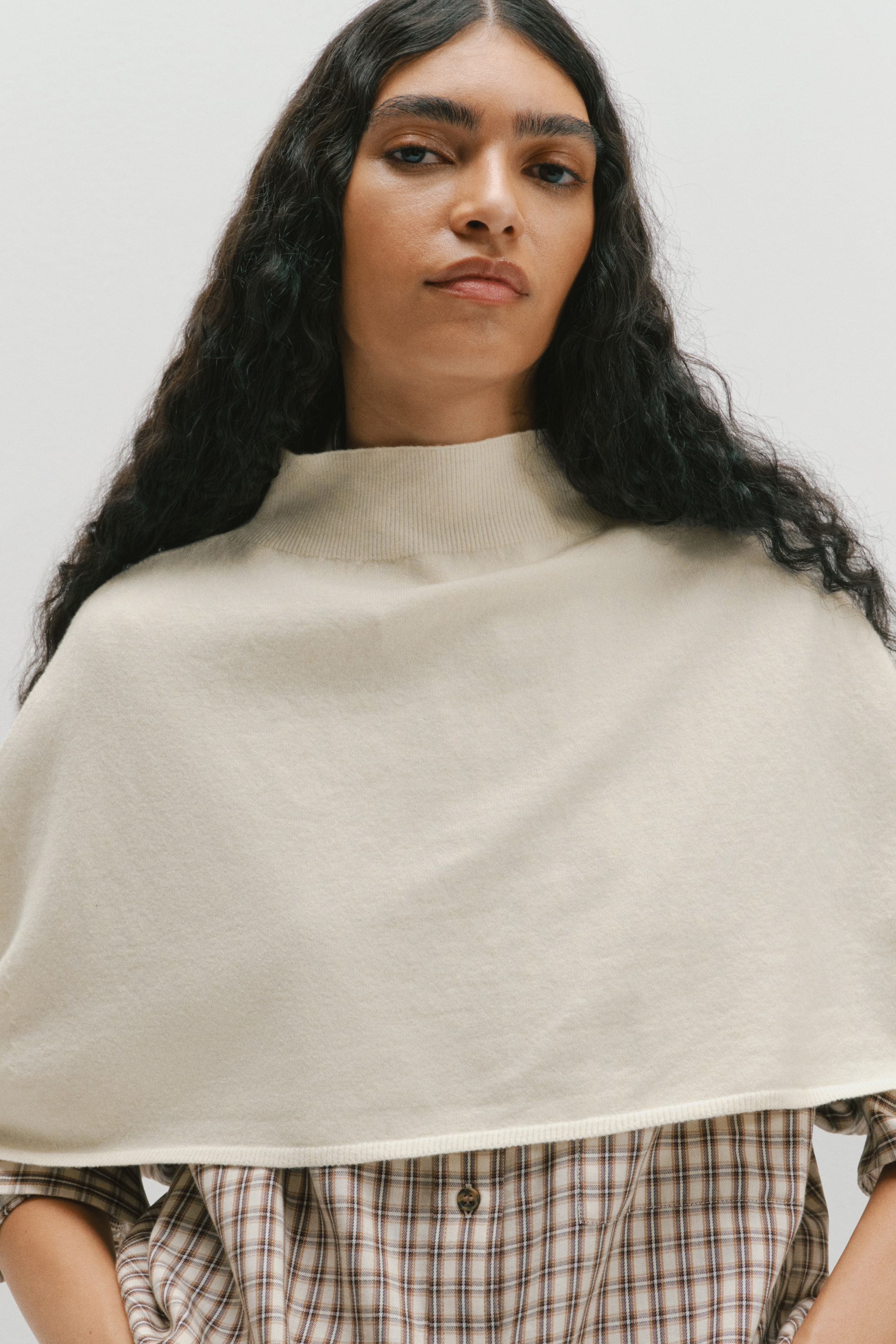 Agrandir l'image: Une cape en maille douce couleur crème est portée par le mannequin, avec un col montant côtelé et un design drapé. La cape est superposée sur une chemise à carreaux boutonnée aux tons neutres.