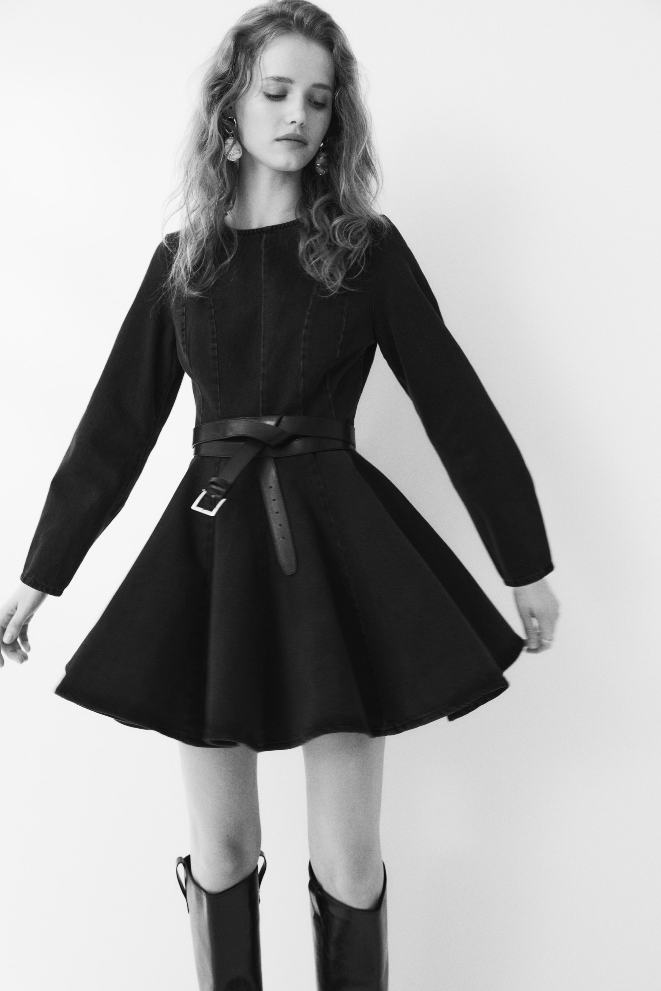 View larger image: The black dress features long sleeves, a round neckline, and a fitted bodice with vertical seams. A dark belt cinches the waist, tying at the front with a visible silver-toned buckle, while its A-line skirt flares out to showcase its volume.