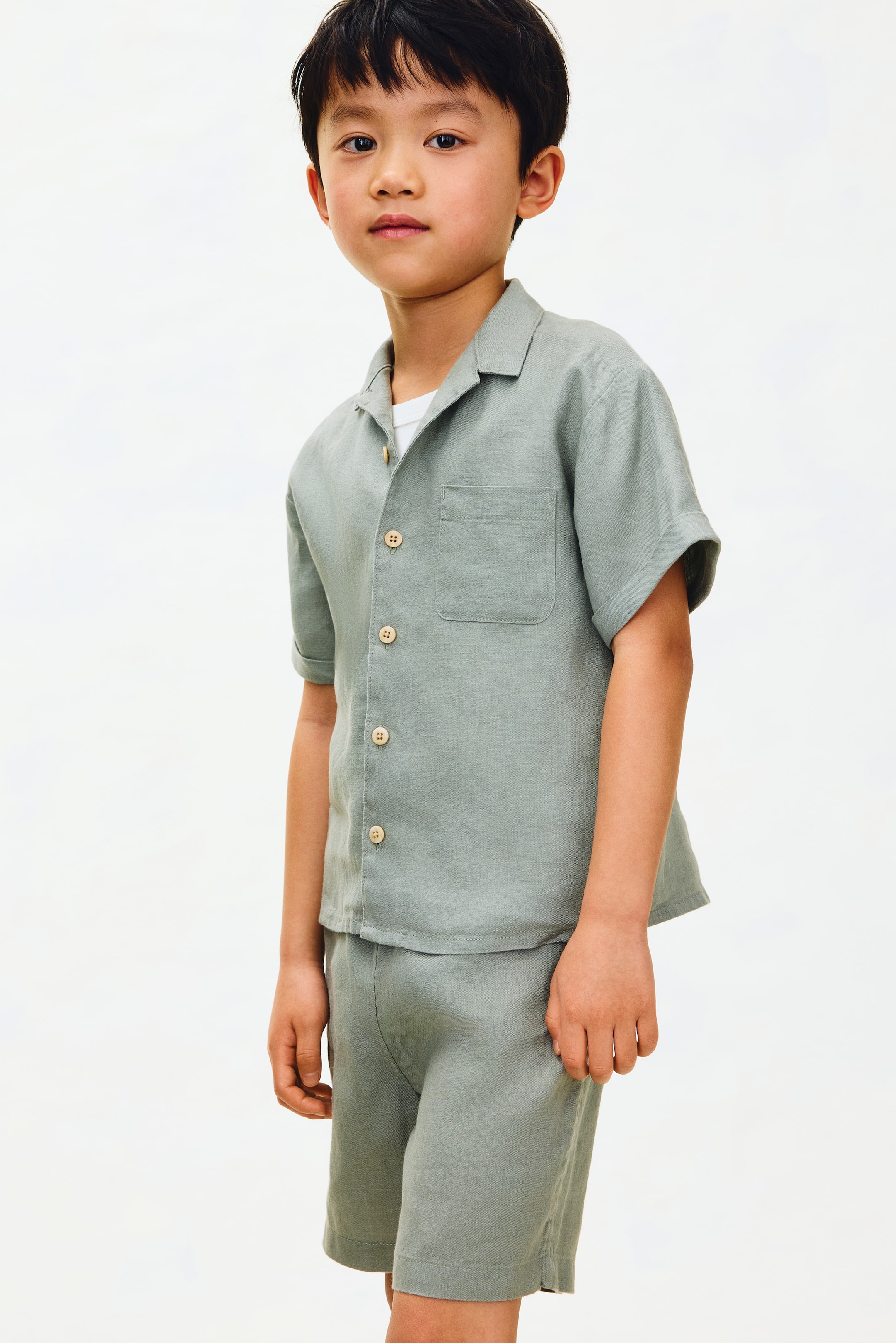 查看大圖: 兒童款質感鼠尾草綠短袖襯衫和配套短褲套裝正在展示中。襯衫採用露營領設計，正面全開扣，搭配淺色圓形鈕扣，左胸設有一個貼袋，內搭白色內衣可見。配套短褲長度至大腿中部，呈現出輕鬆的版型。