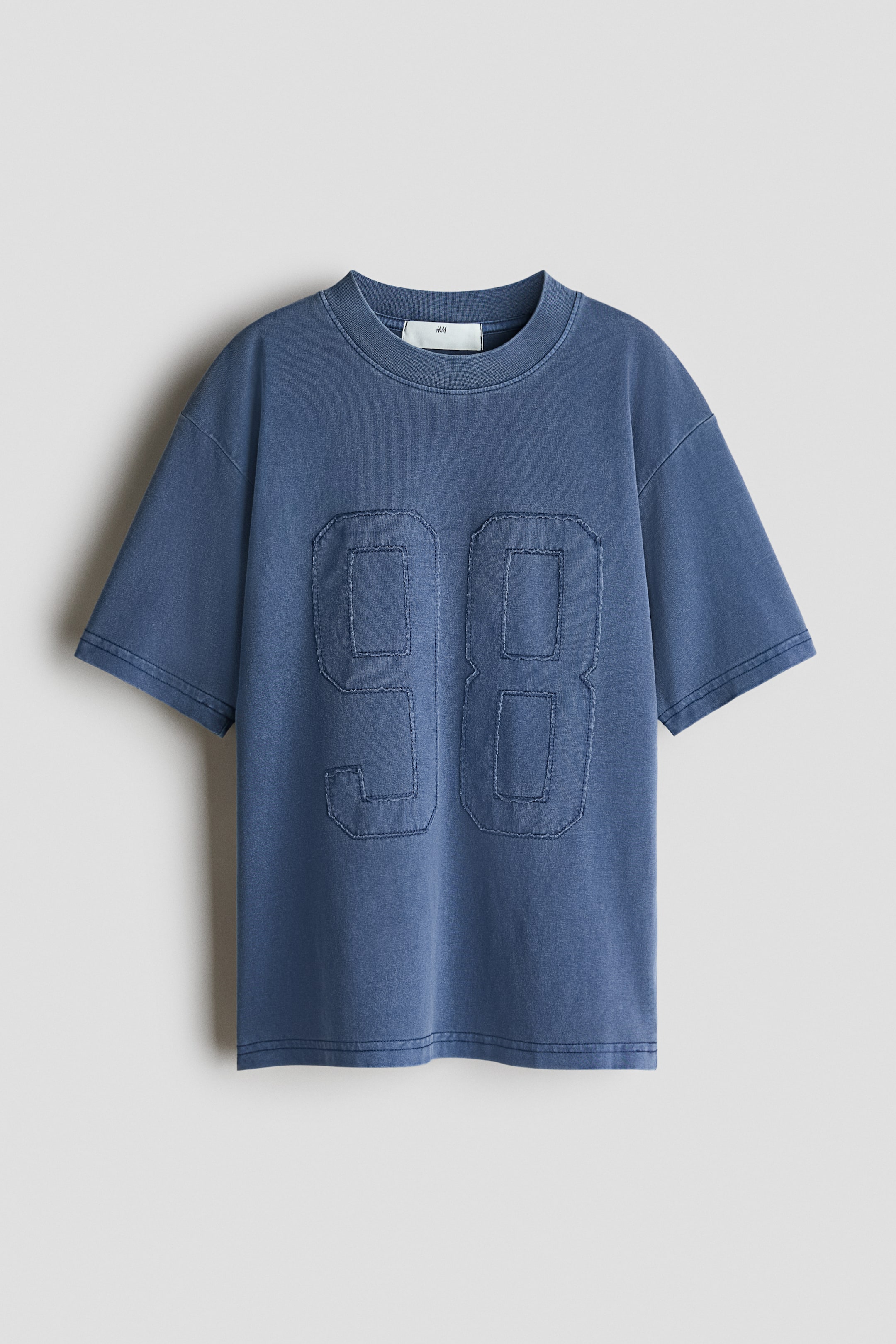 Agrandir l'image: Un t-shirt à coupe ample en jersey de coton doux est présenté de face. Il arbore un col rond bordé de côtes, des épaules légèrement tombantes et un motif imprimé '98' avec des bords usés sur le devant.